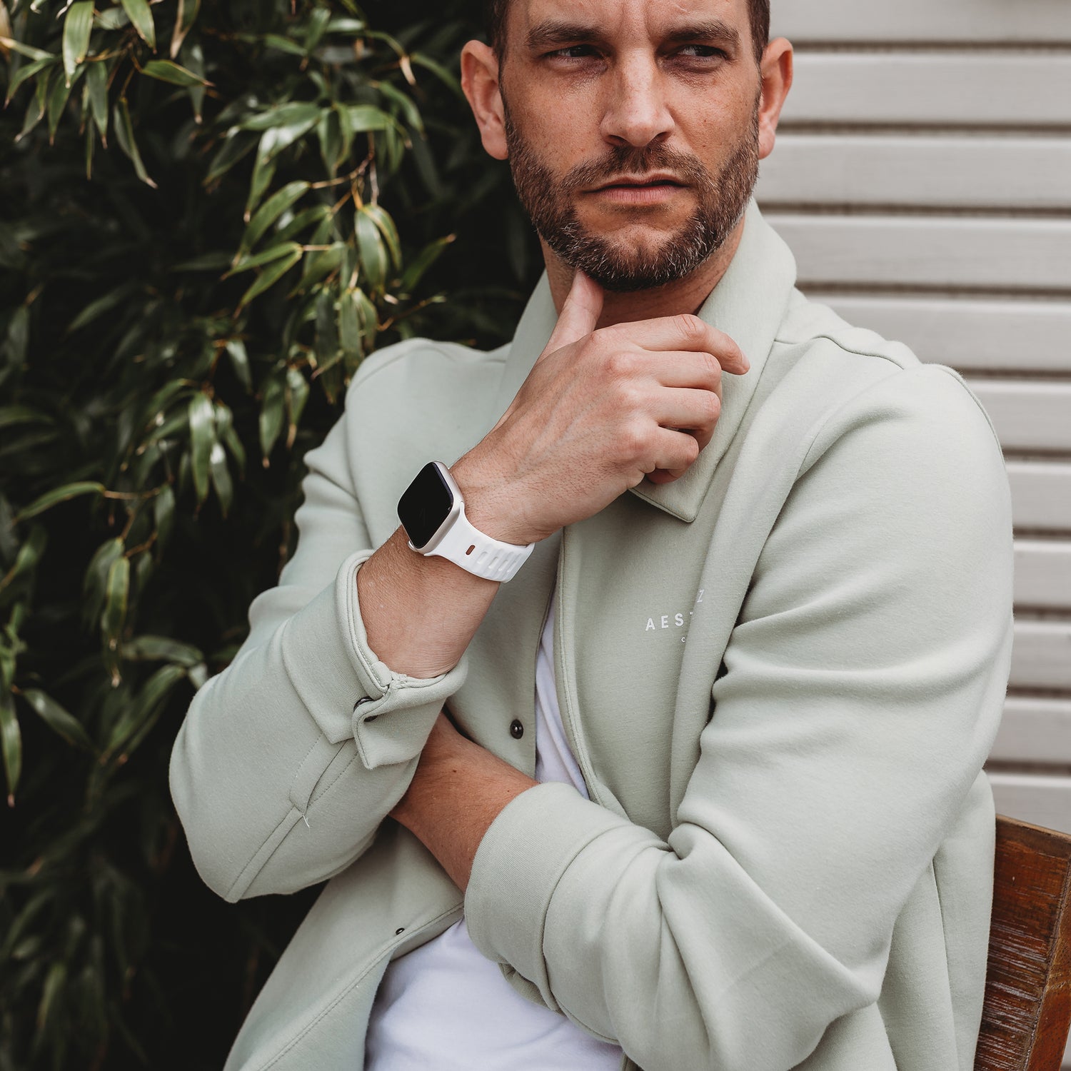 Ein Mann mit gestutztem Bart sitzt im Freien und stützt sein Kinn auf seine Hand. Er trägt eine hellgrüne Jacke, ein weißes T-Shirt und ein Arktisband Apple Watch Silikonarmband „Active“. Im Hintergrund sind grüne Blätter und eine Holzbank zu sehen.