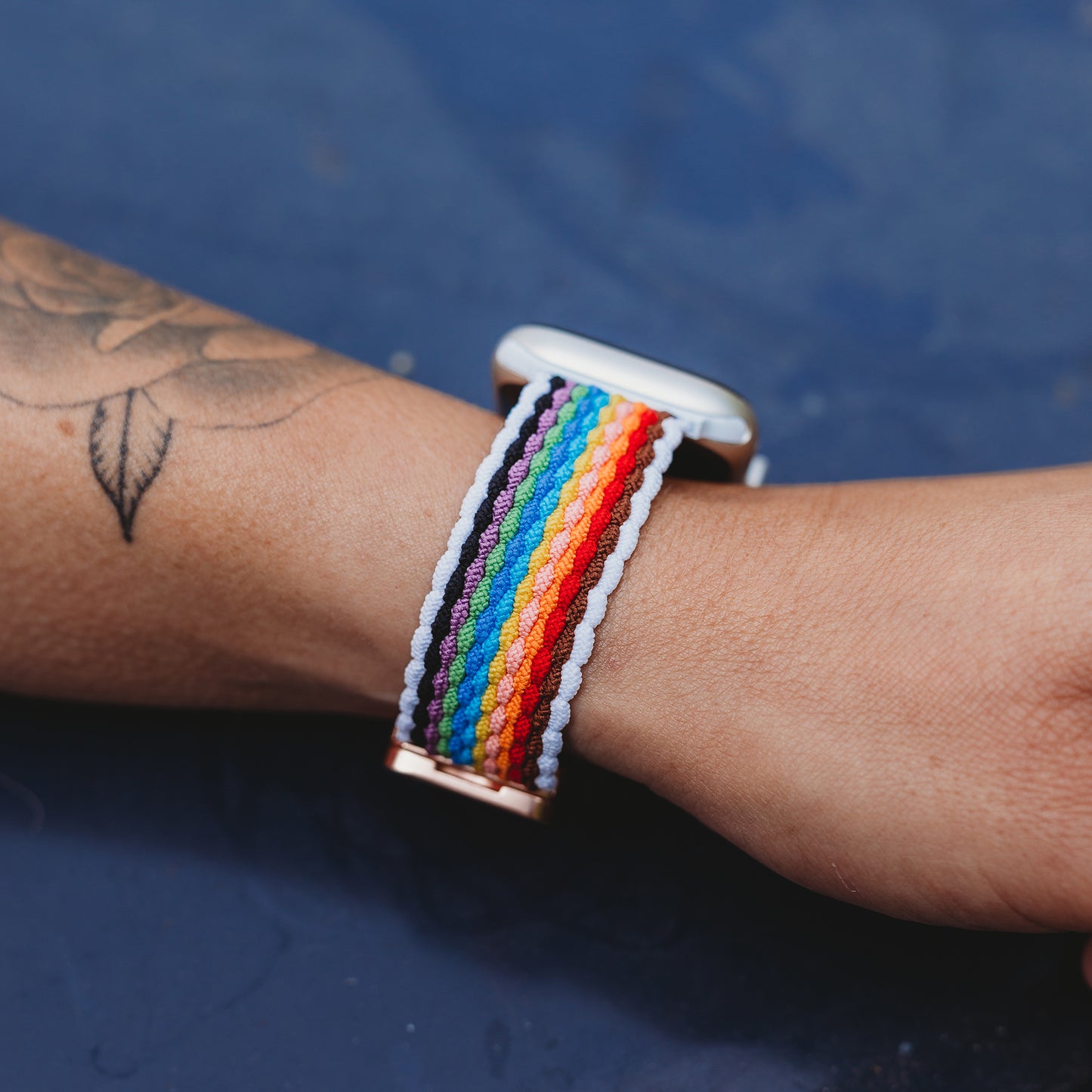 Eine Nahaufnahme eines Handgelenks mit einer Tätowierung, das das arktisband Geflochtene Apple Watch Armband „Casual“ Rainbow Edition mit Regenbogenstreifen, weißen Rändern und magnetischem Metallverschluss vor einem dunkelblauen Hintergrund trägt.