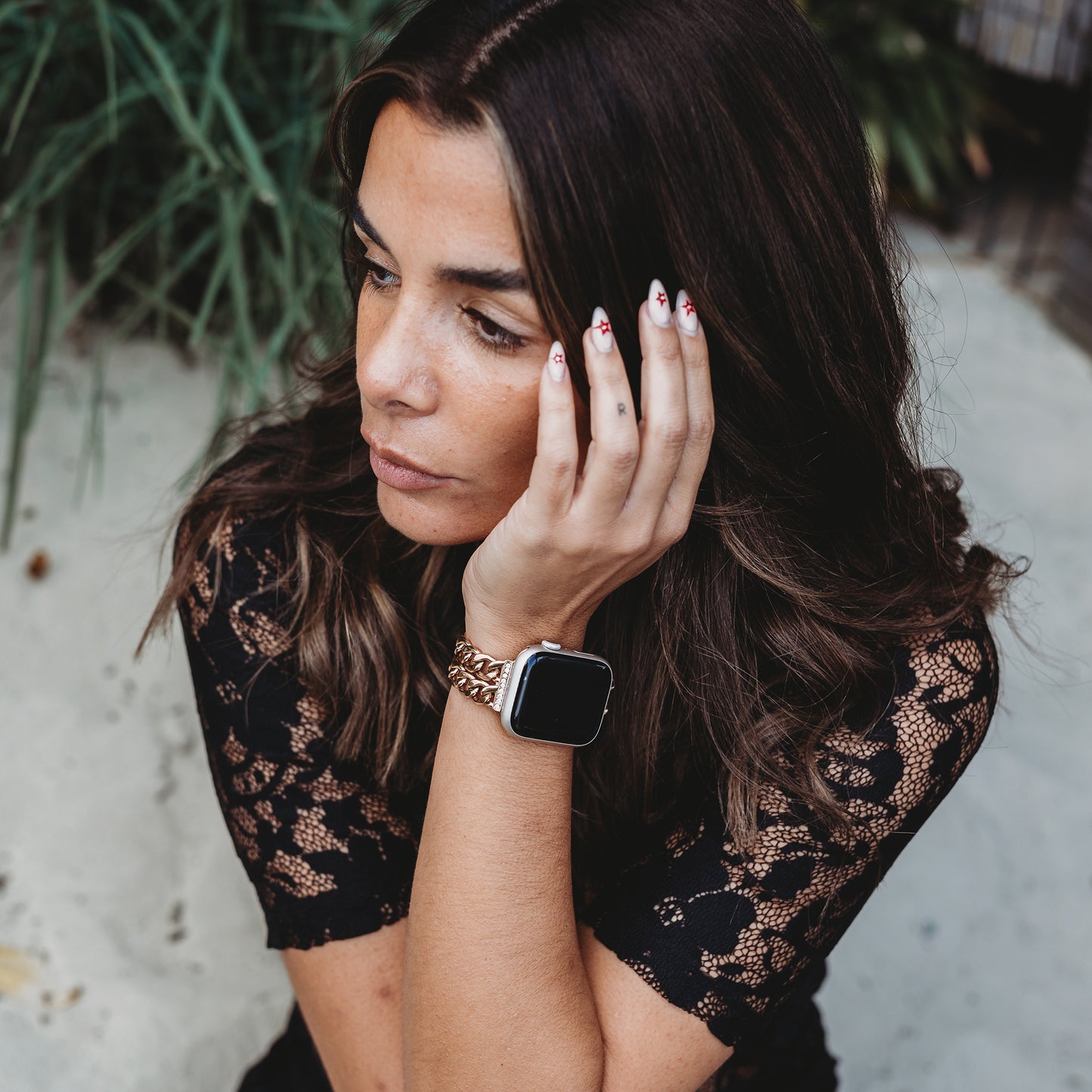 Eine Frau mit langen braunen Haaren und roten Nägeln, die ein schwarzes Spitzentop trägt, stützt ihr Gesicht auf ihre Hand. Sie trägt das arktisband Apple Watch Armband „Princess“ mit Strasssteinchen-Adapter und blickt nachdenklich zur Seite.