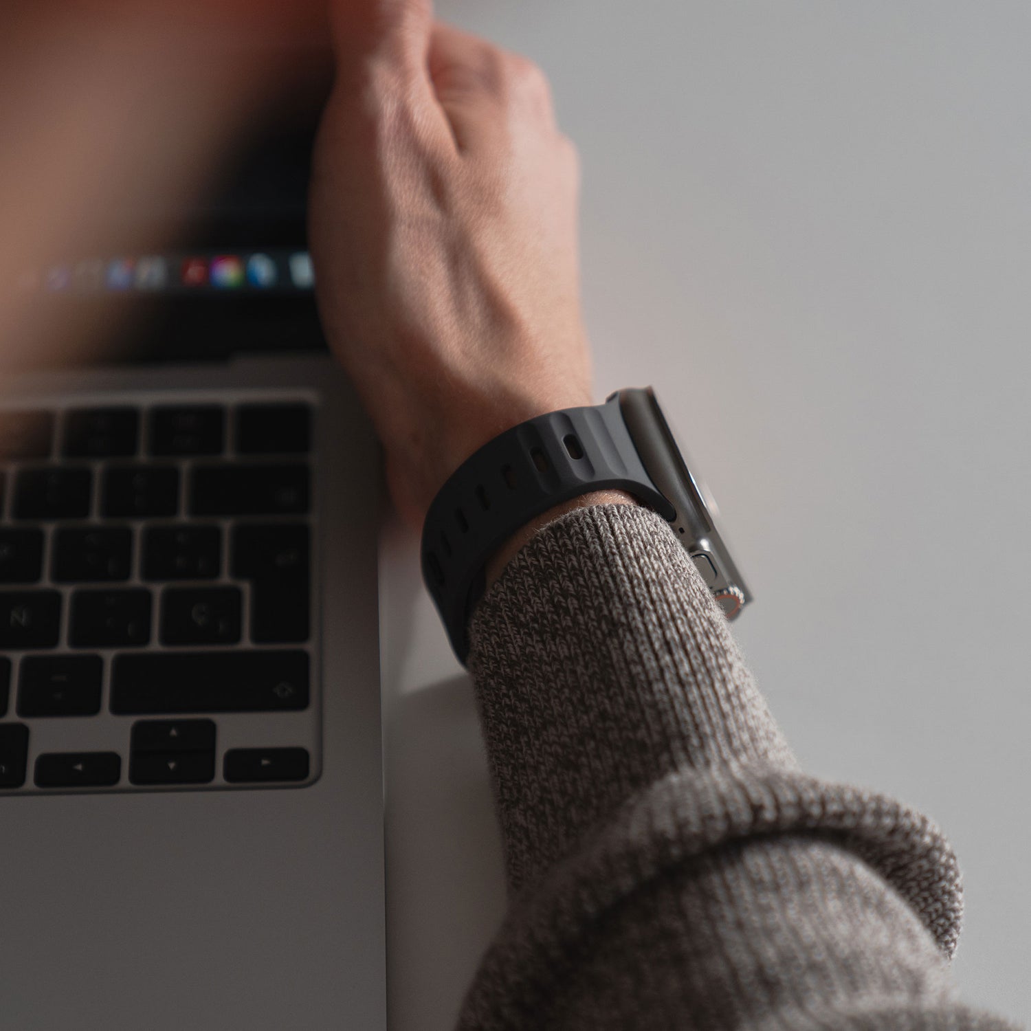 Eine sportlich aussehende Person trägt ein graues Langarmshirt und ein arktisband Apple Watch Silikonarmband „Active“ von arktisband, während sie einen Laptop benutzt und ihre Hand in der Nähe der Tastatur auf einer weißen Oberfläche ablegt.