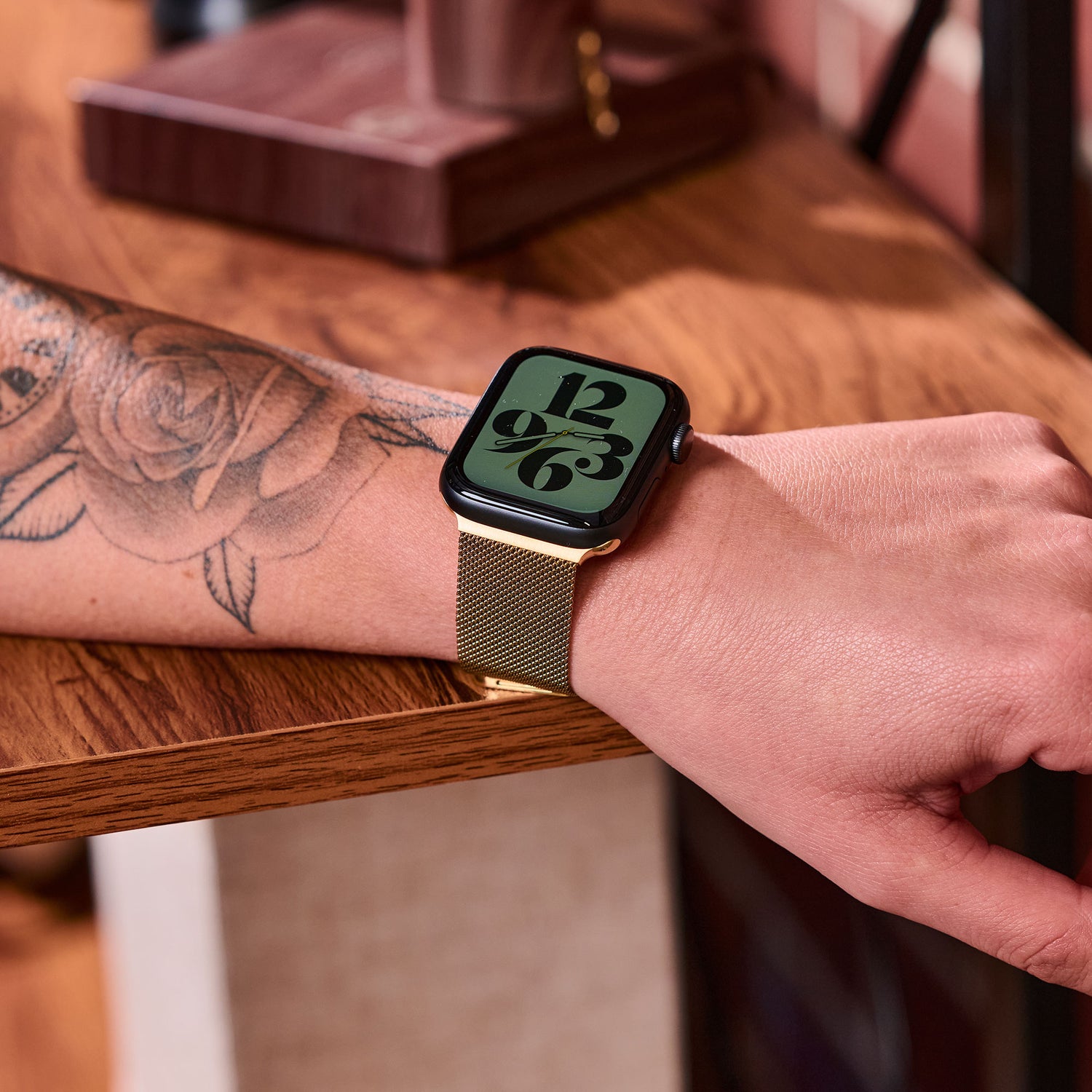 Eine Person mit einem floralen Unterarmtattoo trägt ein goldenes Arktisband Apple Watch Milanaise Loop Armband. Die Uhr zeigt ein großes, markantes digitales Zifferblatt, während der Arm auf einer Holzoberfläche ruht.