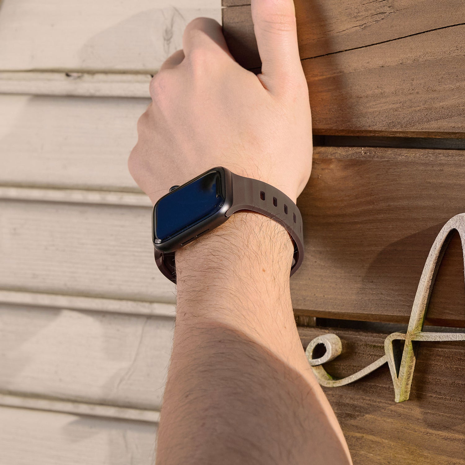 Eine Person hält mit erhobenem linken Arm eine Holzplatte an einer Wand. Sie trägt ein arktisband Apple Watch Silikonarmband „Active“ mit atmungsaktivem Design und ausgeschaltetem Display. Marke: arktisband.