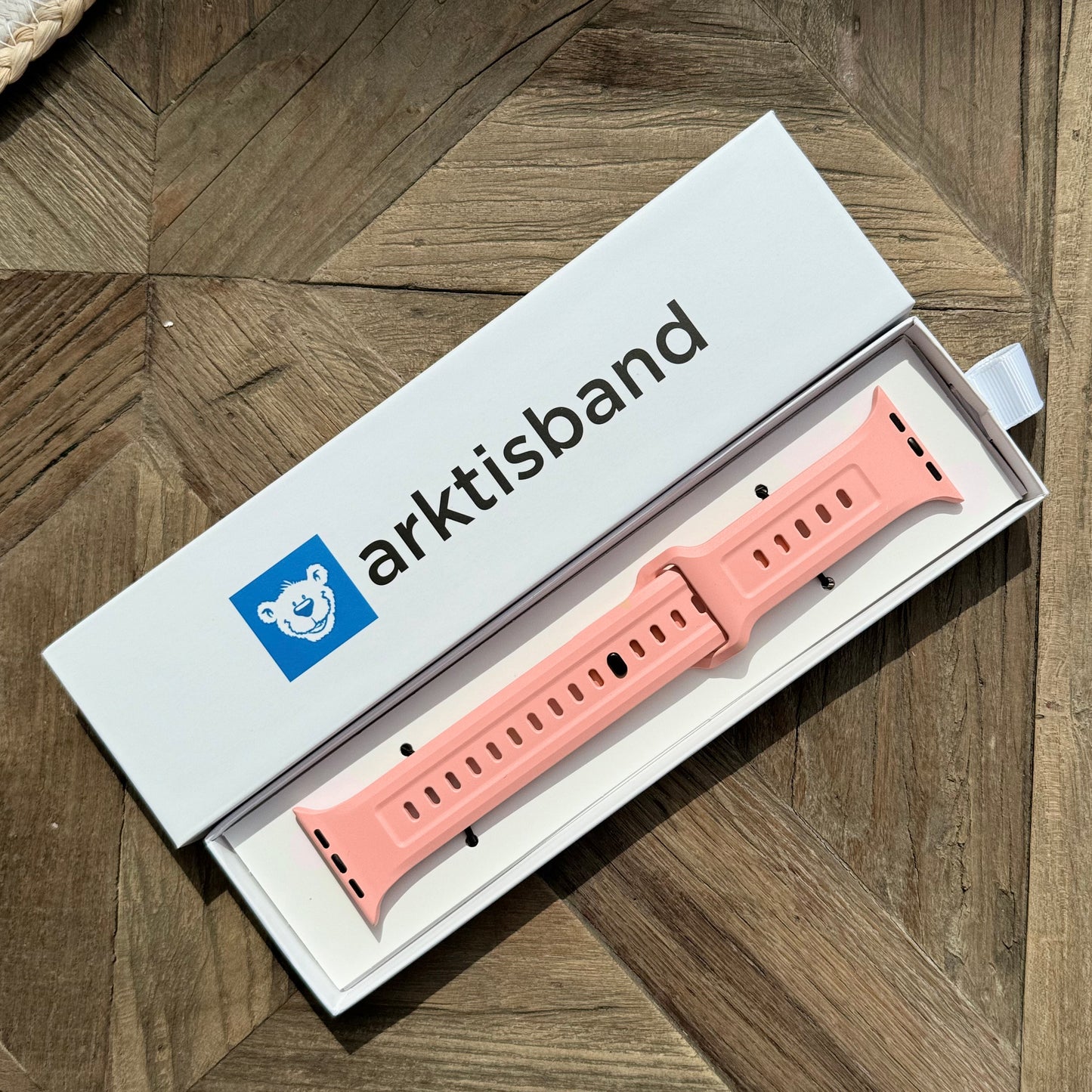 Das arktisband Apple Watch FKM Sportarmband "Sweat", ein pinkes Fluorelastomer-Armband, wird in einer weißen arktisband-Box mit blauem Bären-Logo auf gemusterter Holzoberfläche präsentiert. Das Armband ist, elastisch und Apple Watch kompatibel.