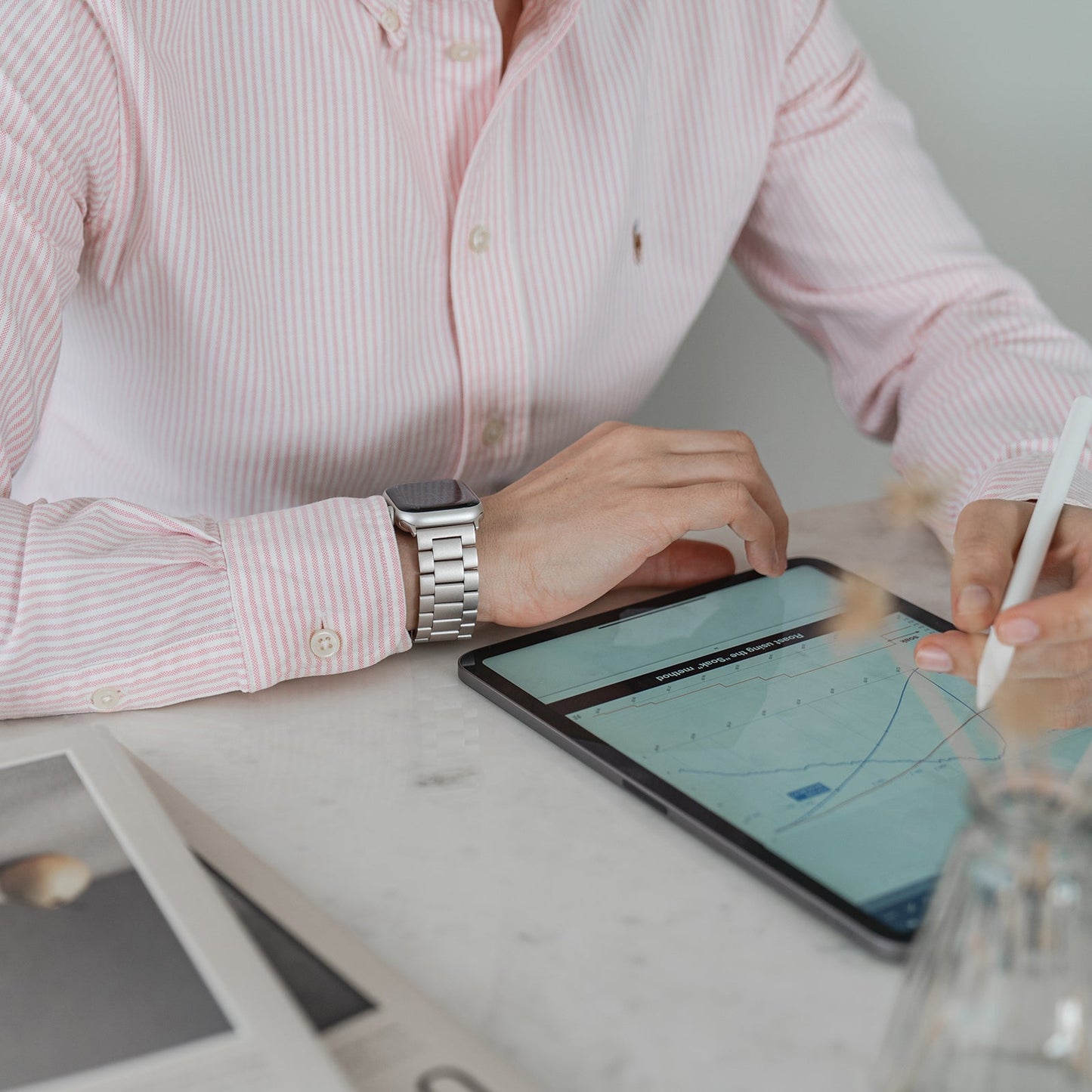 Eine Person in einem rosa-weiß gestreiften Hemd und einem Arktisband-Apple-Watch-Gliederarmband „Arctica“ verwendet einen Stift auf einem Tablet mit einem Diagramm, neben einer Zeitung und einer Glasvase auf dem weißen Tisch.