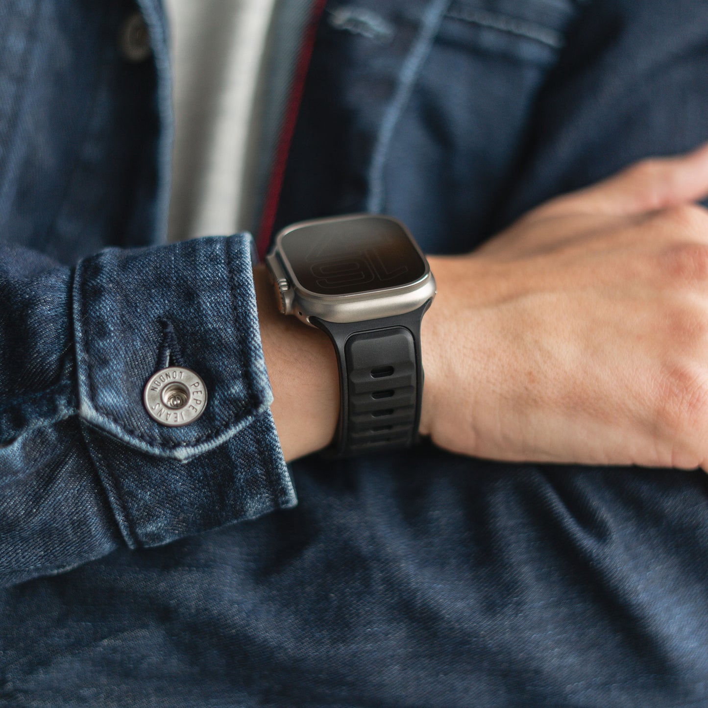 Eine Person in dunkler Jeansjacke und einem arktisband Apple Watch Silikonarmband „Swift“ ruht mit verschränkten Armen auf einer Oberfläche. Das Uhrendisplay ist ausgeschaltet, das schweißabsorbierende, atmungsaktive Armband von arktisband ist sichtbar.