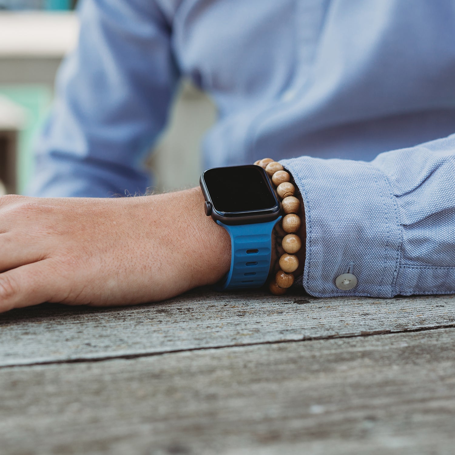 Eine Person, die ein Arktisband Apple Watch Silikonarmband „Active“ und ein Armband aus Holzperlen trägt, ruht mit ihrem Arm auf einem Holztisch und trägt ein hellblaues Hemd mit Knöpfen, das Stil und Atmungsaktivität betont.