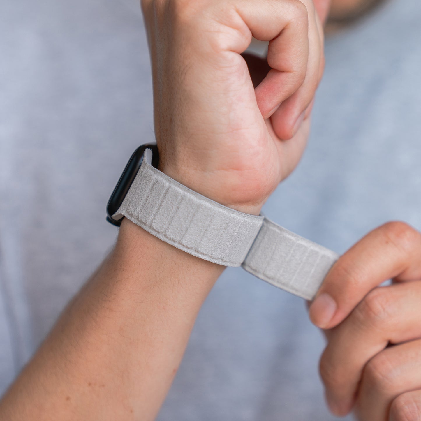 Eine Person passt ein magnetisches Armband aus Alcantara von arktisband Apple Watch in Grau an ihr Handgelenk an und hebt das italienische Design von arktisband vor einem hellen Hintergrund hervor.