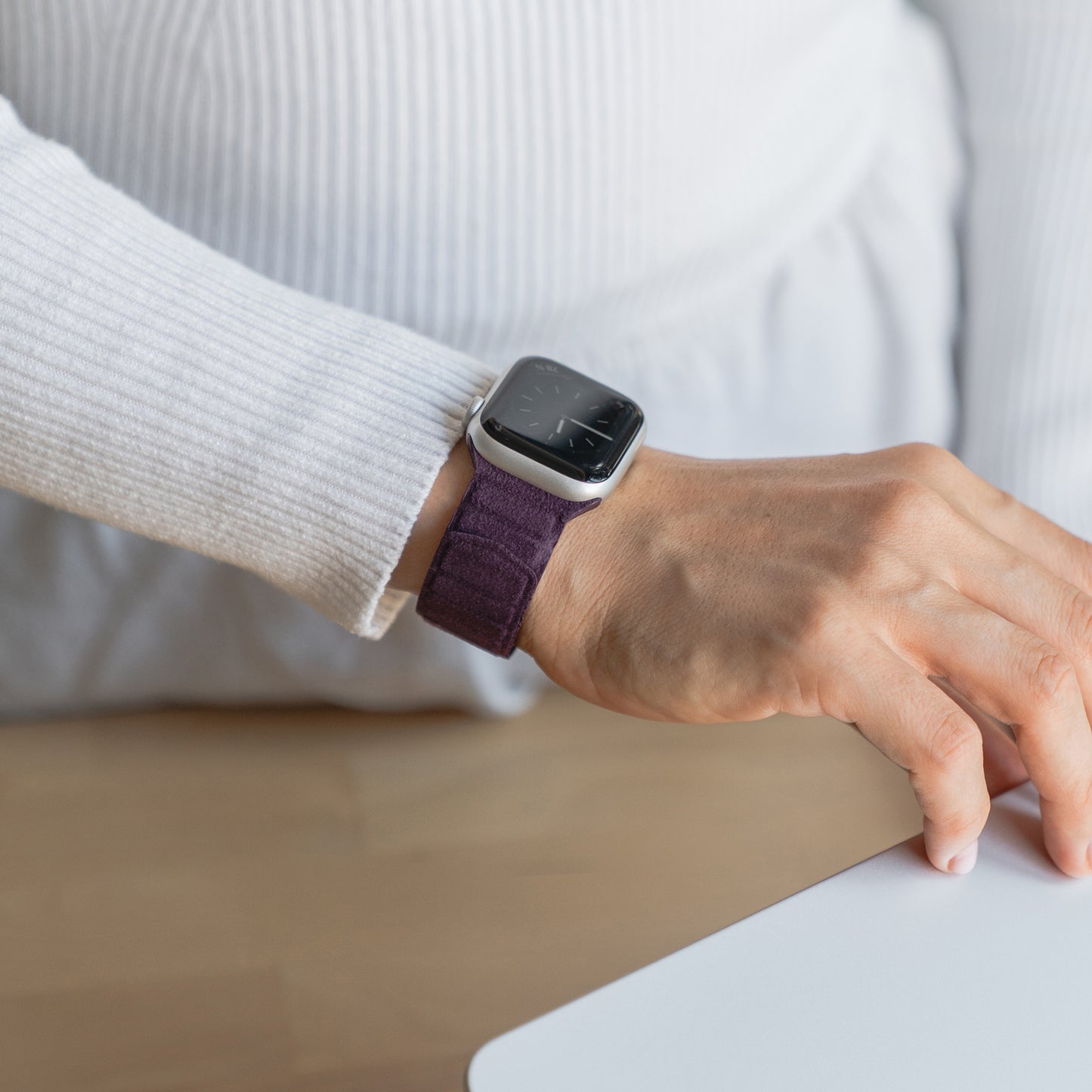 Eine Person, die das arktisband Apple Watch magnetisches Armband aus Alcantara in Lila trägt, stützt ihre Hand auf einen weißen Schreibtisch. Das stylische Band von arktisband ist mit der Apple Watch kompatibel und bringt italienisches Flair in jedes Outfit.