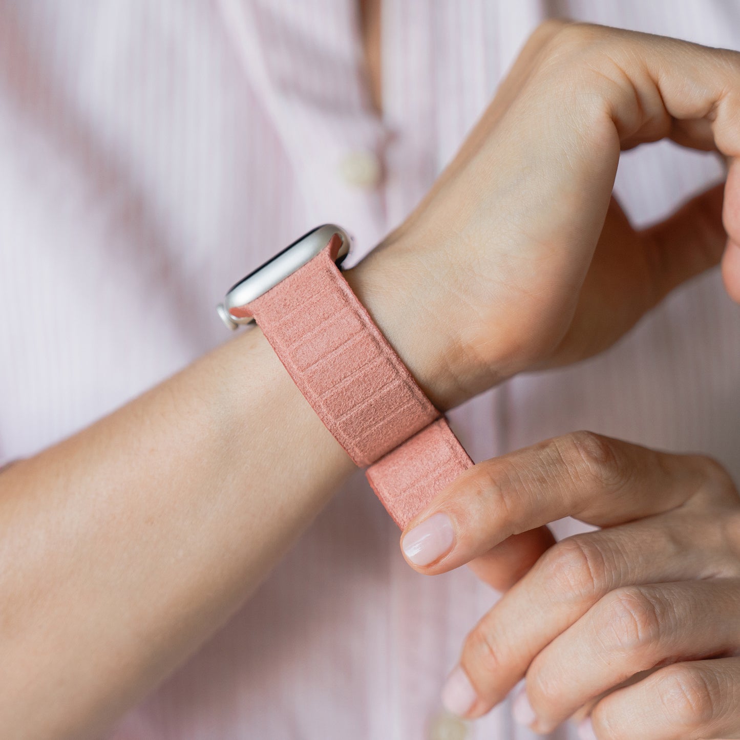 Eine Person passt das arktisband Apple Watch magnetisches Armband aus Alcantara von arktisband an ihr Handgelenk an und trägt ein hellrosa gestreiftes Hemd. Das Band bietet italienisches Design und ist mit der Apple Watch kompatibel.