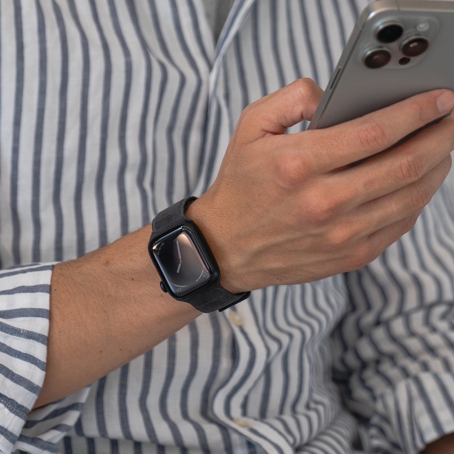 Person im gestreiften Hemd, die ein Smartphone in der Hand hält und ein Arktisband Apple Watch trägt Magnetisches Armband aus Alcantara, das stilvolles italienisches Design zeigt.