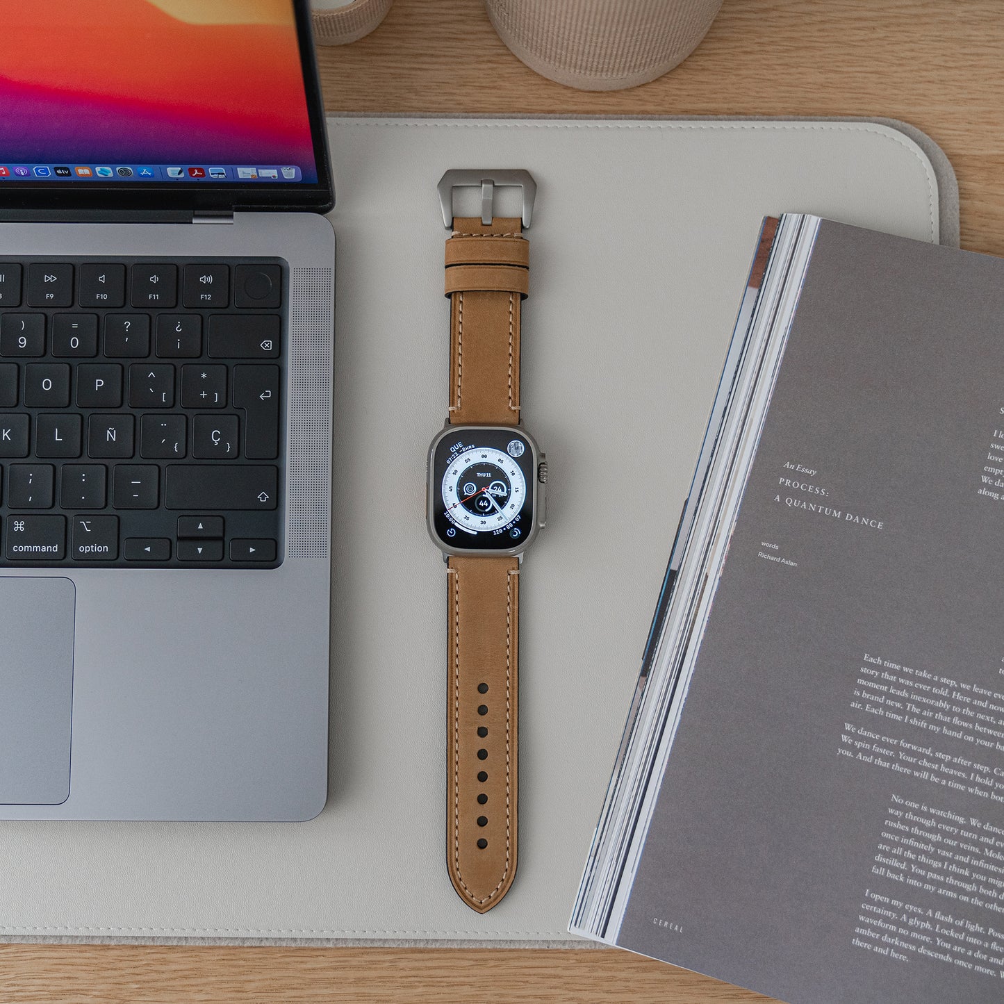 Eine Arktisband Apple Watch Ultra mit dem PALERMO GRANDE Lederarmband liegt auf einer Schreibtischunterlage zwischen einem Laptop und einer offenen Zeitschrift, wobei die Tastatur und das Trackpad des Laptops links teilweise sichtbar sind.