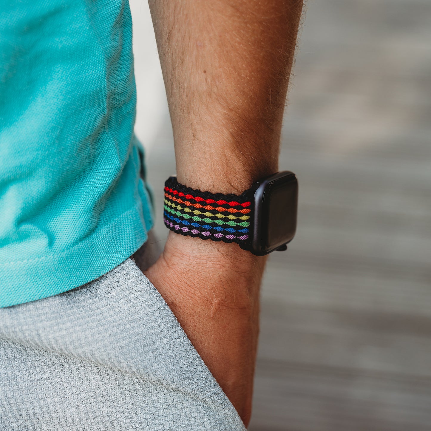 Eine Person in einem blauen Hemd und einer grauen Hose hat die Hand in der Tasche und zeigt das arktisband Geflochtene Apple Watch Armband „Casual“ Rainbow Edition – ein gewebtes Regenbogenband von arktisband – an ihrem Handgelenk.