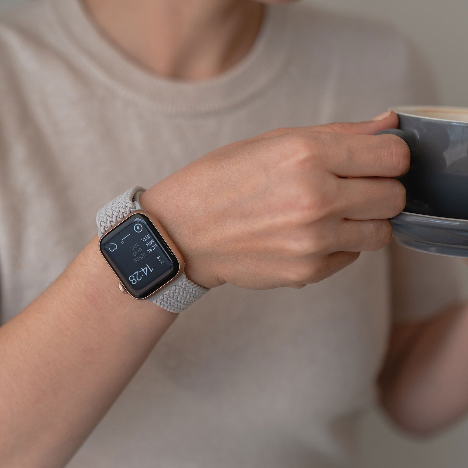 Eine Person trägt ein arktisband Geflochtenes Apple Watch Armband „Casual“ mit Magnetverschluss von arktisband, hält mit beiden Händen eine graue Tasse und ist mit einem hellbeigen Hemd bekleidet.