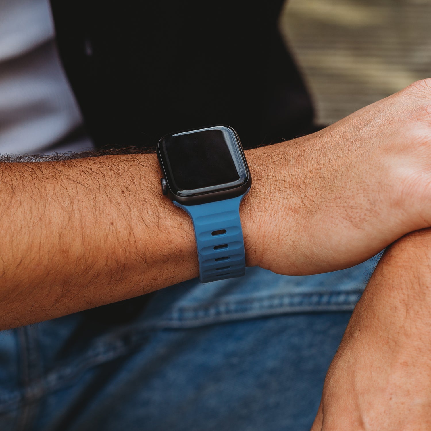 Eine Person trägt ein arktisband Apple Watch Silikonarmband „Active“ in Blau mit ihrer Apple Watch Ultra (Bildschirm ausgeschaltet). Sie trägt ein dunkles Oberteil und blaue Jeans und legt den Arm auf ihr Bein, was die Atmungsaktivität des Armbands betont.
