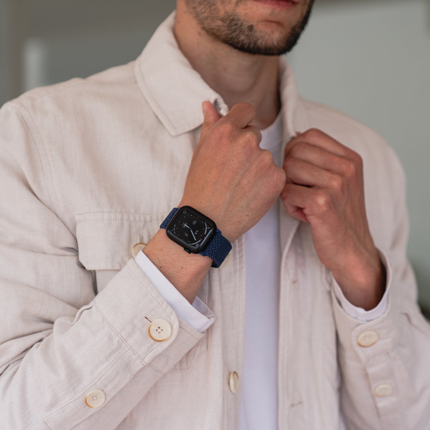 Ein Mann richtet eine hellbeige Jacke und trägt an seinem linken Handgelenk ein von Arktisband geflochtenes Apple Watch Armband „Casual“ mit Magnetverschluss in Dunkelblau.