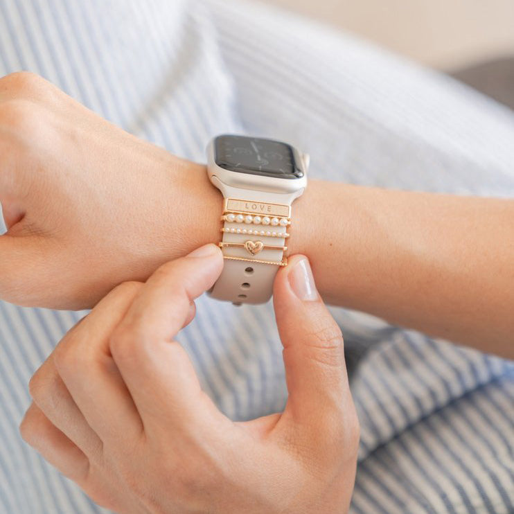 Apple Watch mit weißem Silkonarmband und aufgesteckten, goldfarbenen Schmuckelementen.