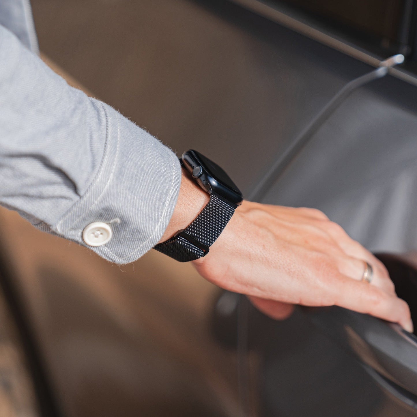 Eine Person in einem grauen Langarmshirt und einem Arktisband Apple Watch Milanaise Loop Armband öffnet eine Autotür. Sie trägt außerdem einen silbernen Ring am Ringfinger.