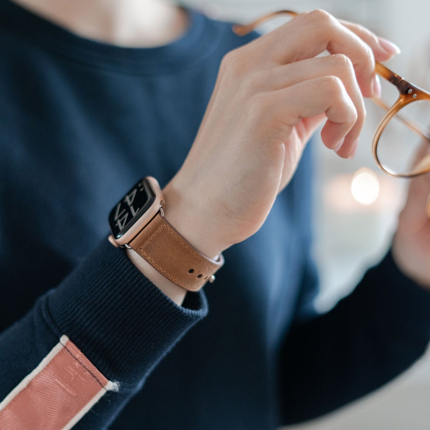 Eine Person in einem marineblauen Pullover mit rosa Streifen rückt eine braune Brille zurecht, während sie das arktisband Apple Watch Lederarmband „Novus“ von arktisband trägt, das maximalen Tragekomfort und einen minimalistischen Look an ihrem Handgelenk bietet.