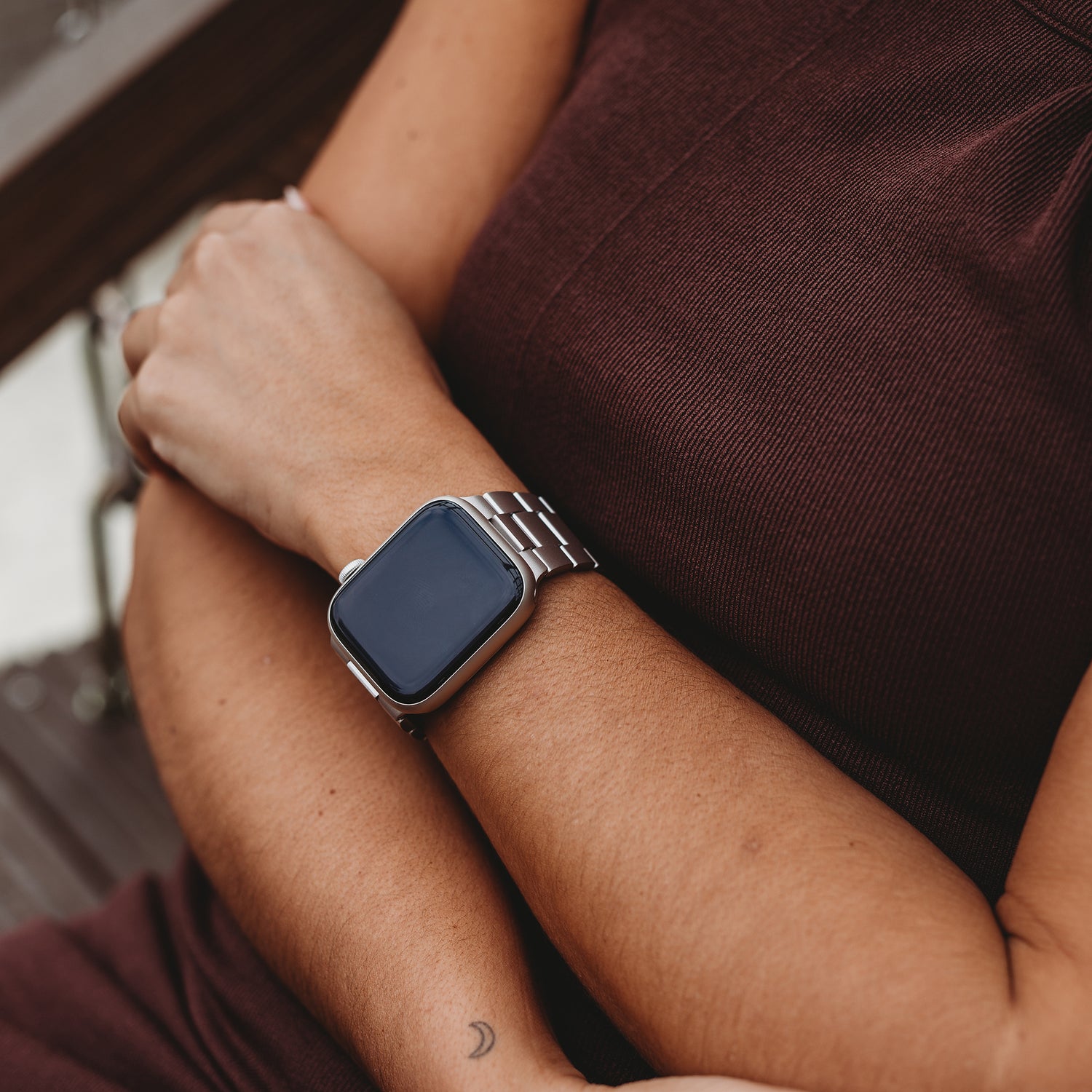 Eine Person in einem braunen ärmellosen Kleid mit verschränkten Armen trägt ein arktisband Apple Watch Gliederarmband „Prime“ von arktisband. Auf ihrem linken Unterarm ist ein Halbmond-Tattoo zu sehen, das Display der Smartwatch ist leer.