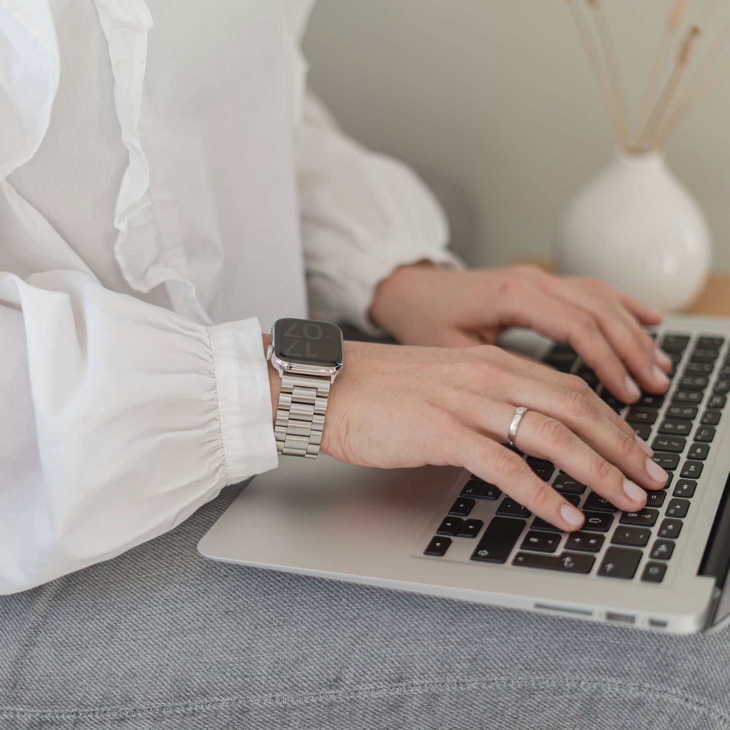 Eine Person trägt das Arktisband Apple Watch Gliederarmband „Arctica“ und einen Ring und tippt auf einem Laptop, bekleidet mit einer weißen Bluse und Bluejeans, im Hintergrund steht eine weiße Vase.