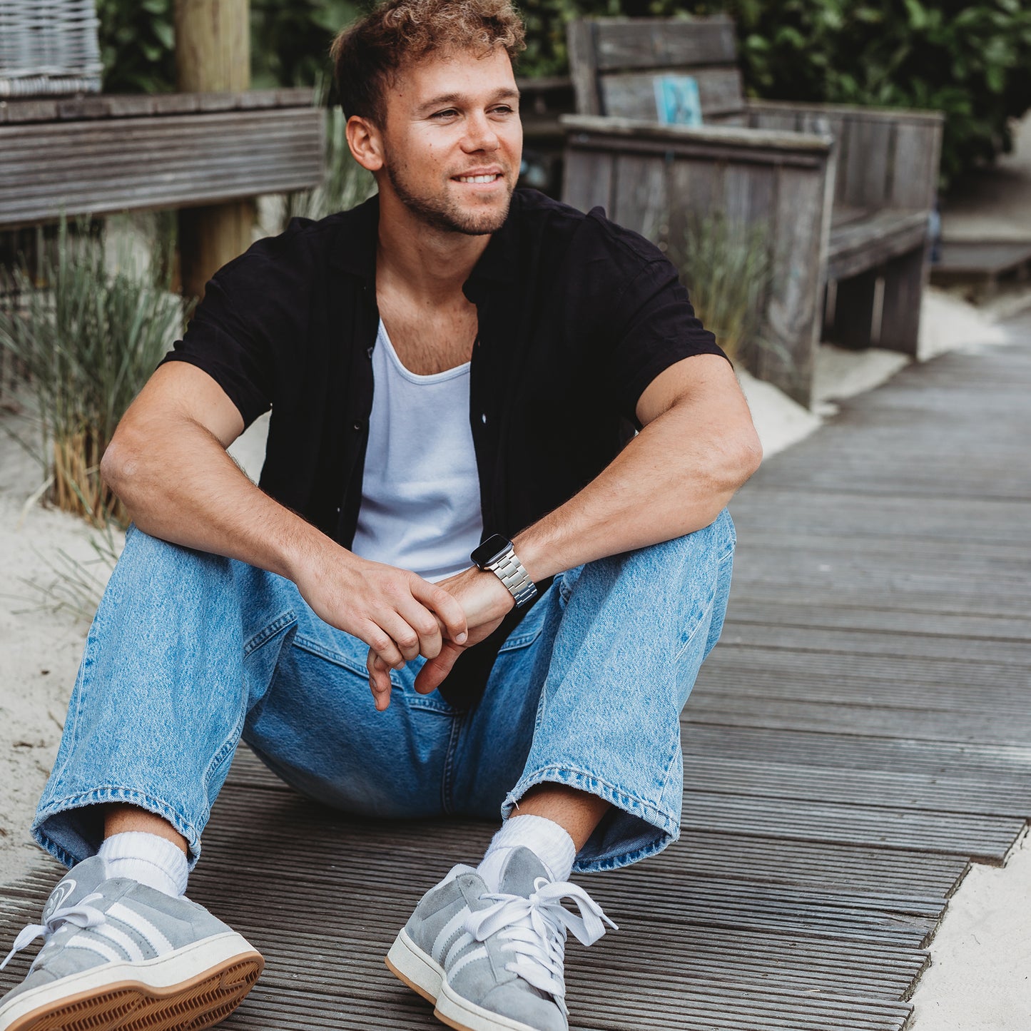 Ein Mann mit kurzen Haaren und Bart sitzt auf einem Holzsteg am Sandstrand. Er trägt ein schwarzes Hemd über einem weißen T-Shirt, blaue Jeans, graue Turnschuhe und ein Arktisband Apple Watch Gliederarmband „Prime“ und lächelt, während er zur Seite schaut.