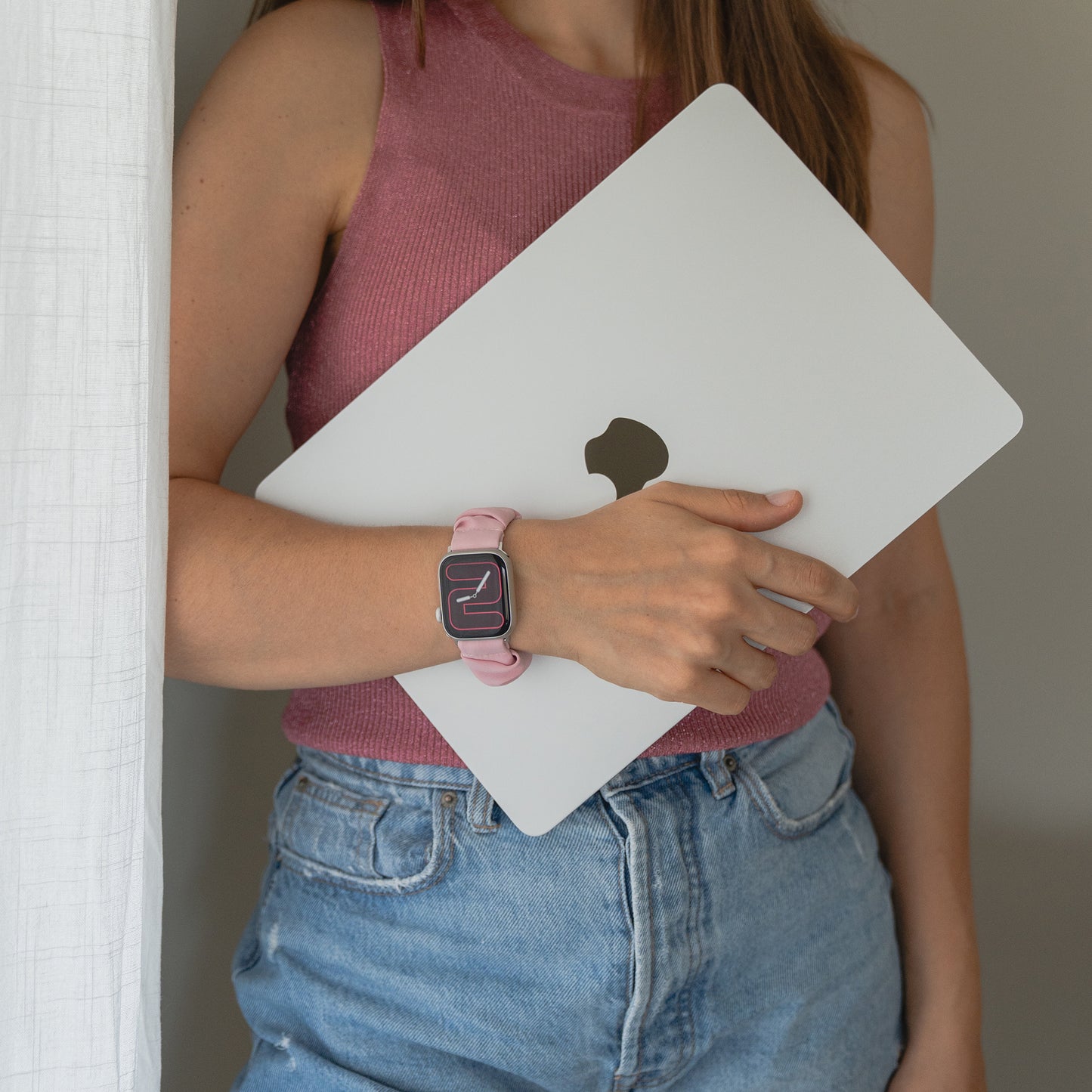 Eine Frau in einem rosa Top und blauen Jeans hält einen geschlossenen Laptop und trägt das arktisband Apple Watch Scrunchie Armband "Elanora" von arktisband.