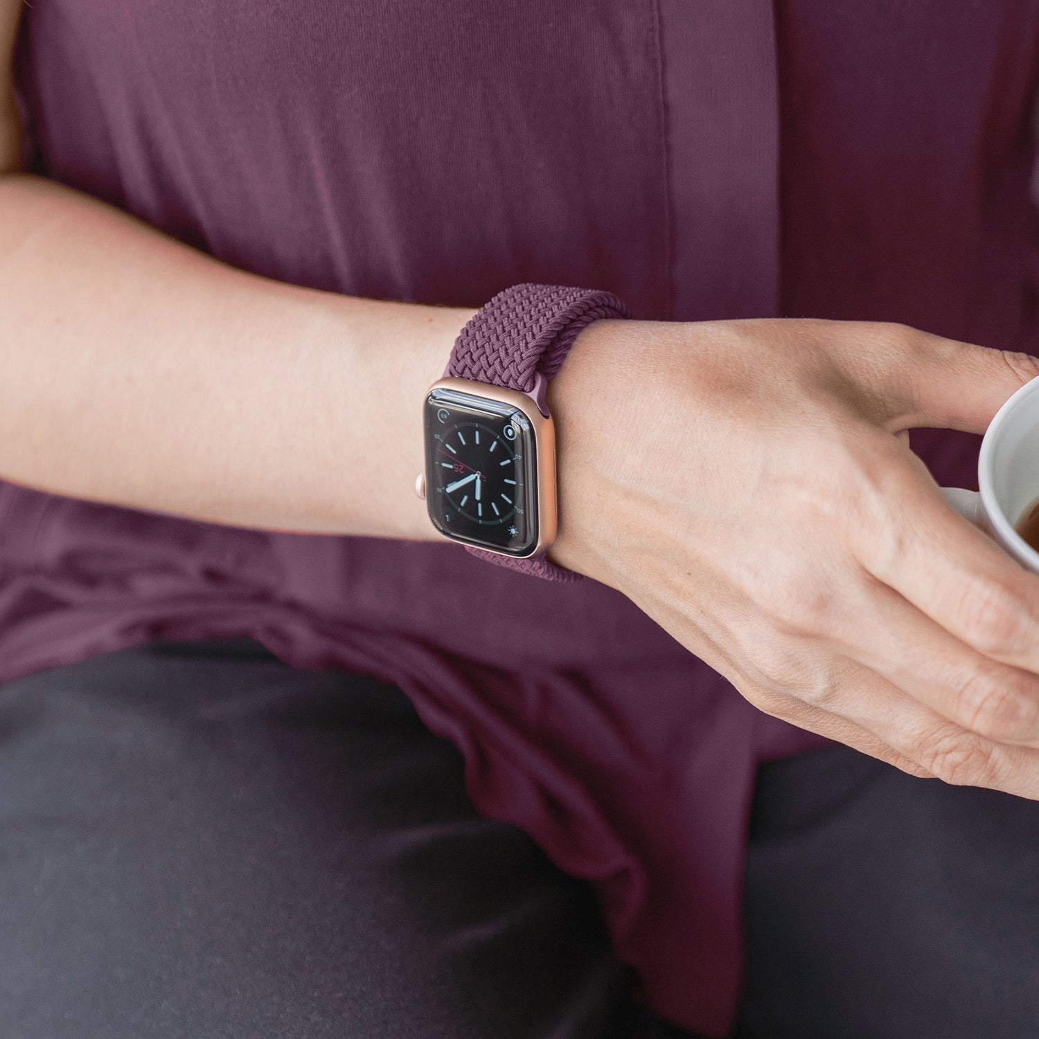Jemand, der ein geflochtenes Apple Watch Armband „Casual“ mit Magnetverschluss trägt, hält eine kleine Tasse in Lila und Schwarz.