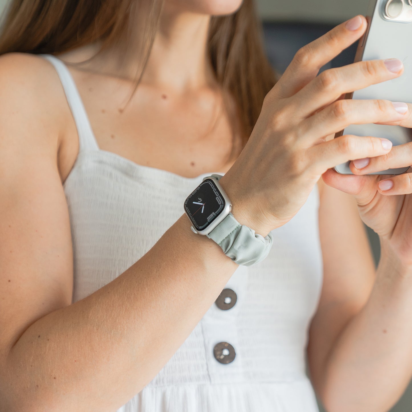 Frau in einem weißen Kleid, die ein Smartphone benutzt, trägt das arktisband Apple Watch Scrunchie Armband "Elanora" von arktisband - ein helles, elastisches Band für mühelosen Stil.