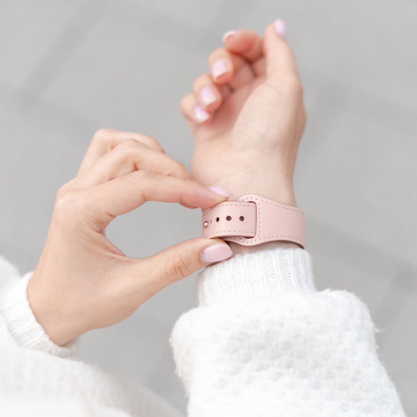 Eine Person im weißen Pullover befestigt das arktisband Apple Watch Lederarmband „Novus“ mit hellrosa Armband am Handgelenk. Ihre hellrosa manikürten Nägel ergänzen den minimalistischen Look vor einem weichen, unscharfen grauen Hintergrund.