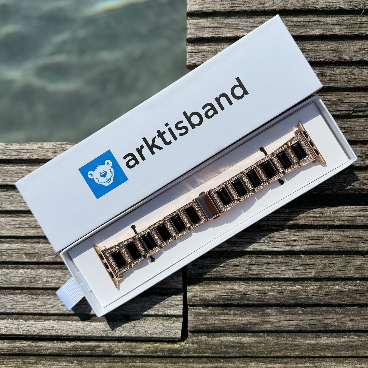 Eine weiße rechteckige Box mit der Aufschrift „arktisband“ und einem blauen Bärenlogo enthält das arktisband Apple Watch Gliederarmband „Senorita“ in Roségold mit schwarzen Gliedern, das auf einer Holzoberfläche am Wasser ruht.