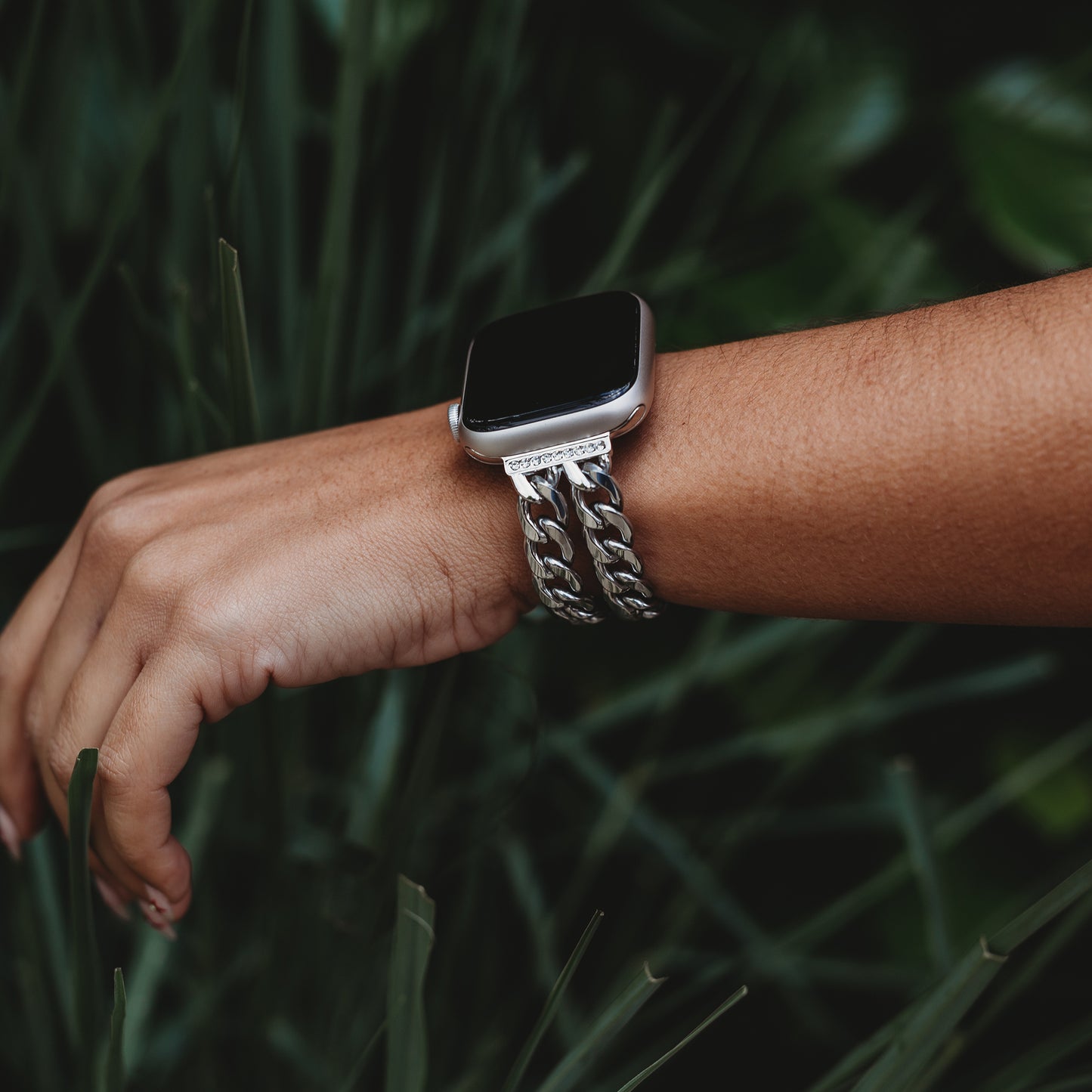 Eine Nahaufnahme des Arms einer Person, die das Arktisband Apple Watch Armband „Princess“ mit einer glanzvollen Gliederstruktur und einem dicken silbernen Kettengliedband trägt, umgeben von grünen Blättern und Pflanzen im Hintergrund.