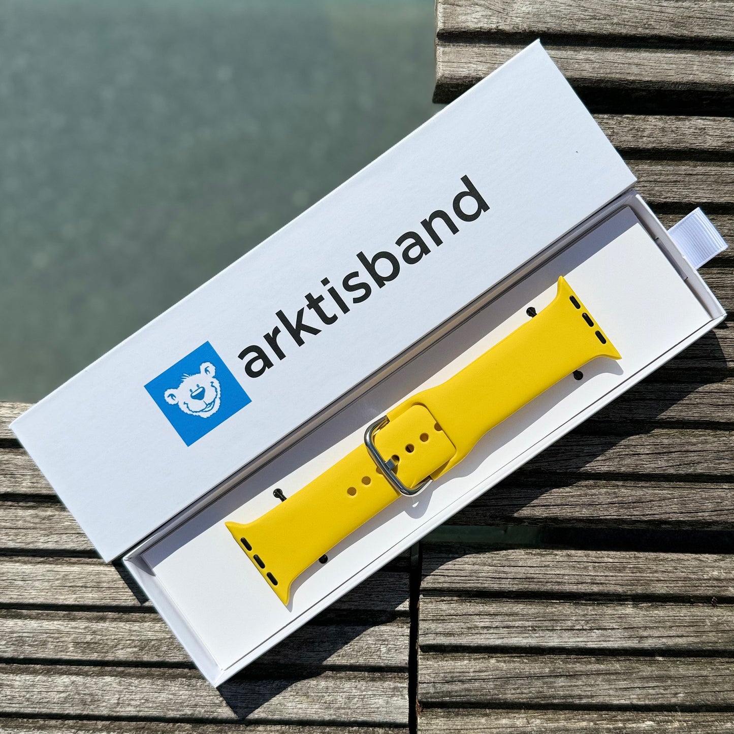 Das arktisband Apple Watch Silikonarmband „Aqua“ wird mit einer silbernen Schnalle in einer weißen arktisband-Box mit blauem Bärenlogo auf einer Holzoberfläche am Wasser gezeigt.