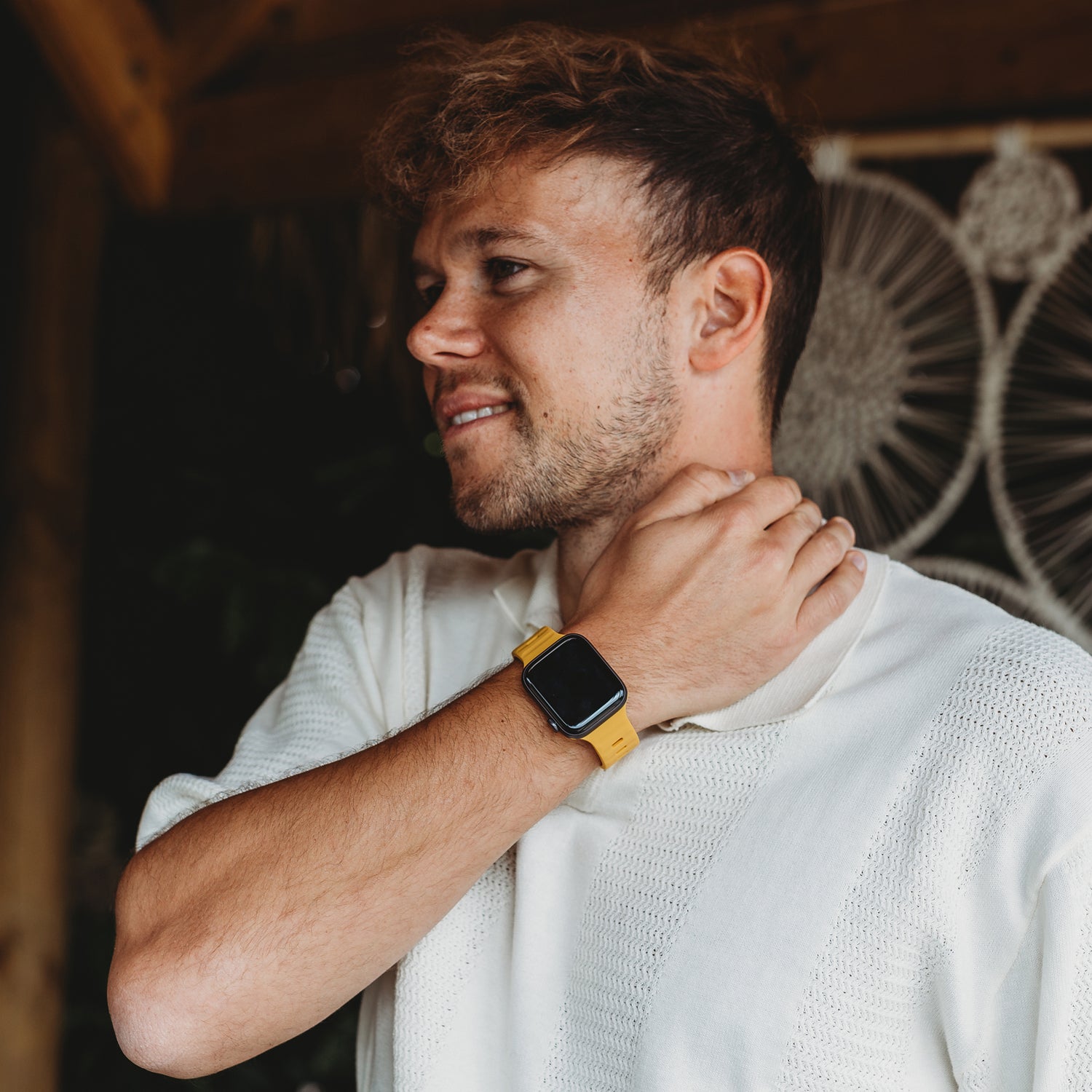 Ein Mann in einem weißen strukturierten Hemd und einem Arktisband Apple Watch Silikonarmband „Active“ von Arktisband lächelt drinnen und berührt seinen Hals mit einem gewebten Wandbehang im Hintergrund.