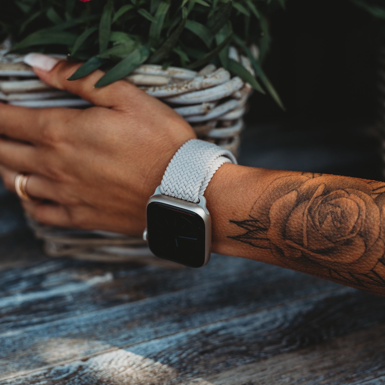 Ein tätowierter Arm trägt ein geflochtenes Apple Watch Armband „Casual“ mit Magnetverschluss von arktisband und hält einen geflochtenen Korb mit grünen Blättern auf einem rustikalen Holztisch.