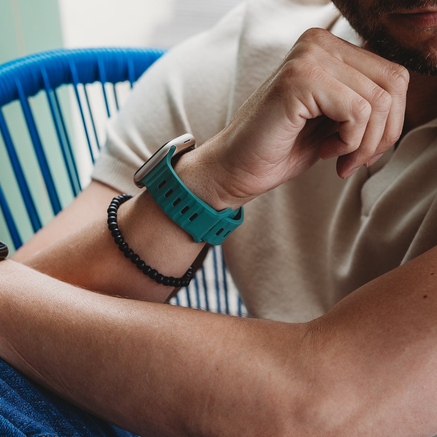 Eine Person mit angewinkeltem Arm trägt das arktisband Apple Watch Silikonarmband "Active" (blaugrünes, atmungsaktives Design) und ein schwarzes Perlenarmband. Sie trägt ein kurzärmeliges helles Hemd und sitzt auf einem blauen Stuhl.