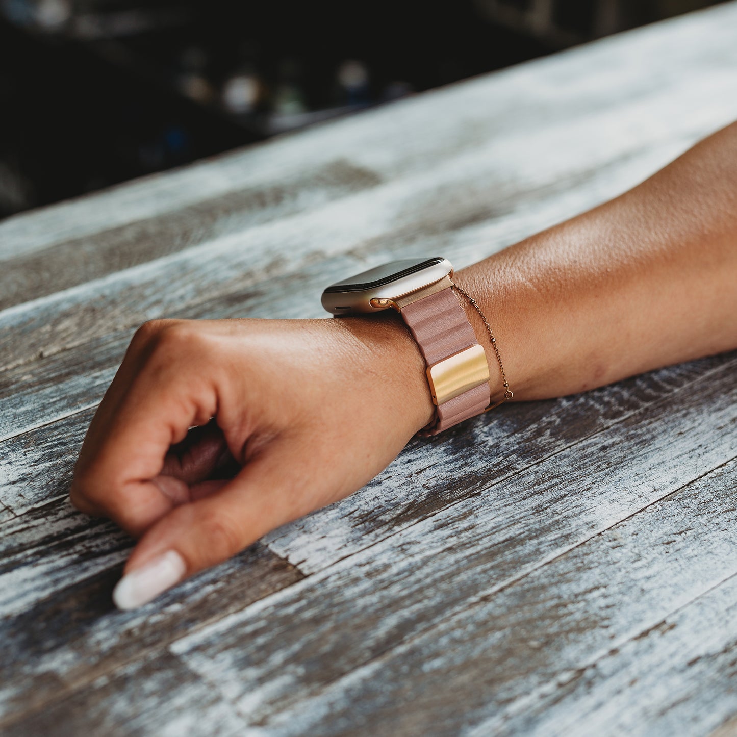 Eine Person mit manikürten Nägeln und hellbrauner Haut ruht mit ihrem linken Arm auf einer Holzoberfläche. Sie trägt das arktisband Kunstlederarmband „Gustino“ für die Apple Watch von arktisband, ein zartgoldenes Metallicband mit Magnetverschluss.