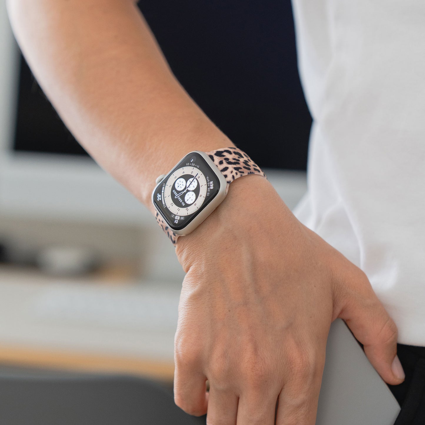 Jemand trägt eine arktisband EDITION für Apple Watch von arktisband mit einem Band mit Leopardenmuster und hält ein graues Objekt vor einem unscharfen Hintergrund.