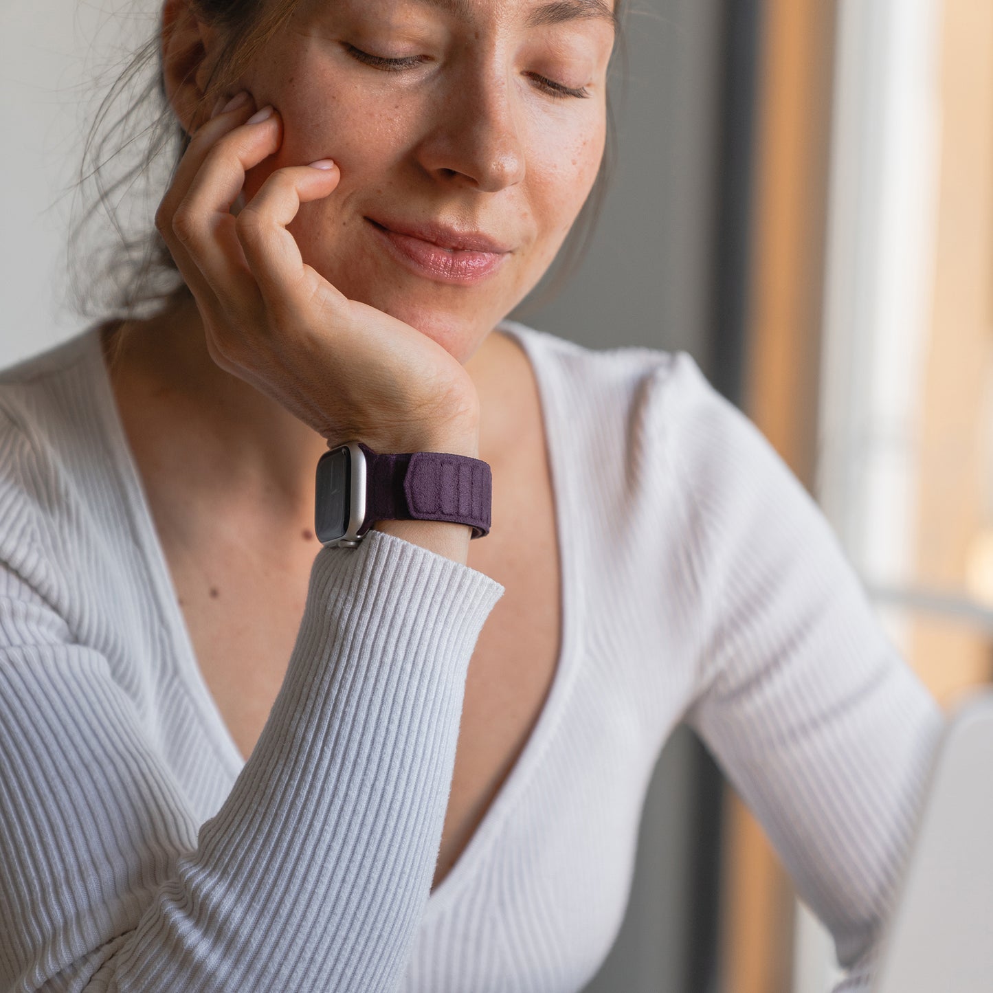 Eine Frau mit geschlossenen Augen lächelt sanft und stützt ihr Kinn auf ihre Hand, während sie das elegante Arktisband Apple Watch magnetisches Armband aus Alcantara trägt.