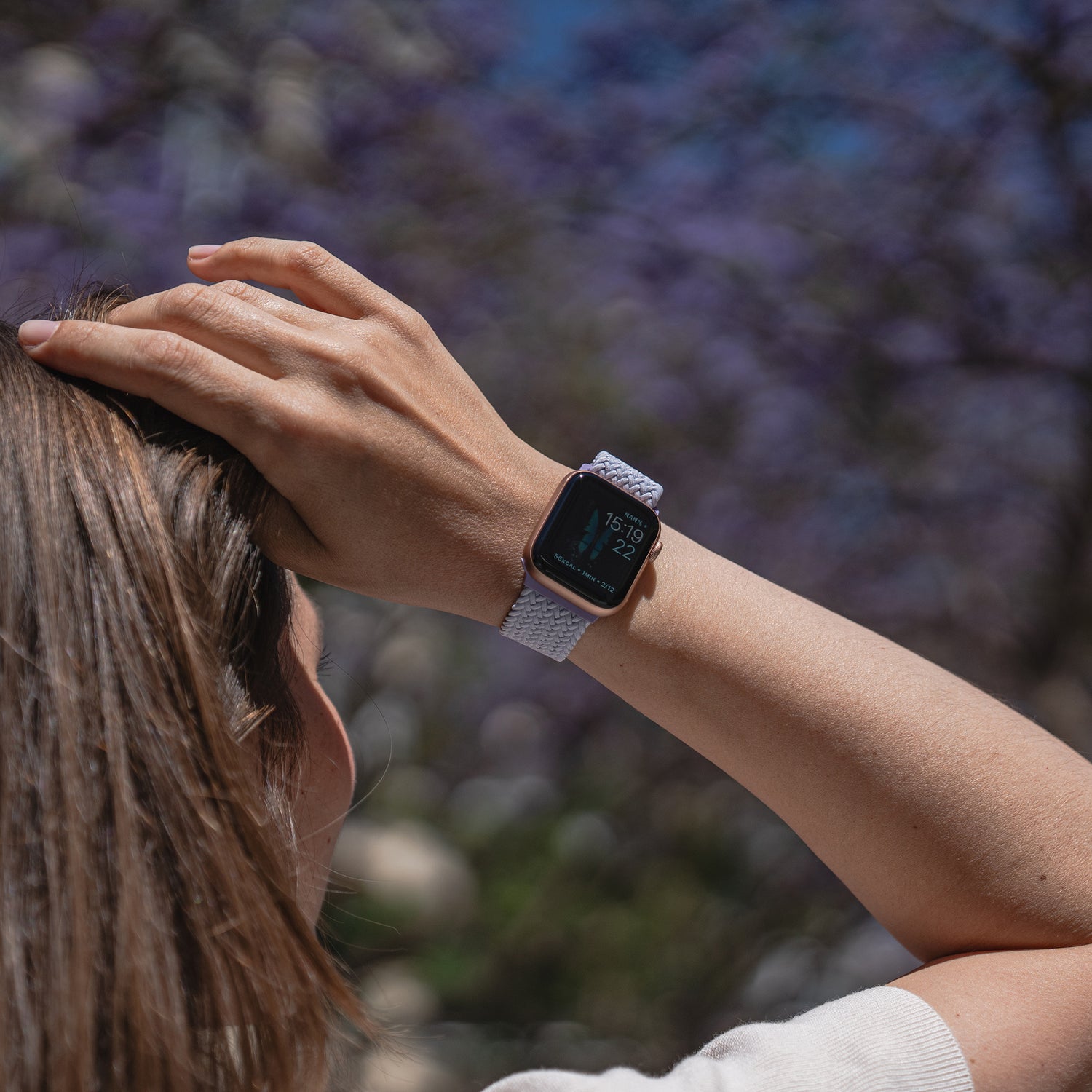 Eine Frau berührt im Freien ihren Kopf. Sie trägt ein geflochtenes Apple Watch Armband „Casual“ mit Magnetverschluss von arktisband, im Hintergrund sind verschwommene lila Blumen zu sehen.