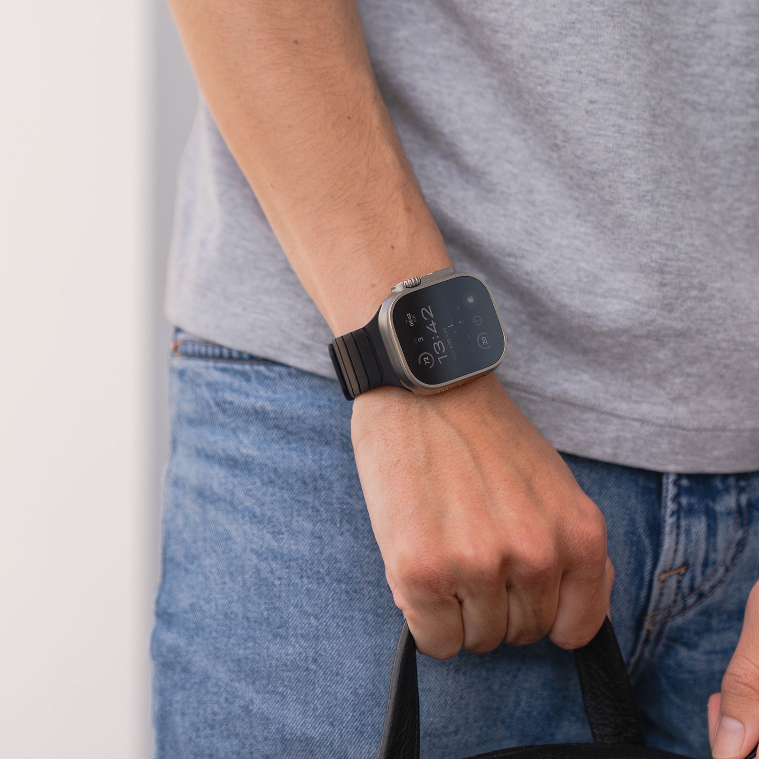Person trägt ein graues T-Shirt und ein schwarzes Arktisband-Magnetarmband „MagFlow“ für die Apple Watch und hält eine schwarze Tasche an ihrer Seite.