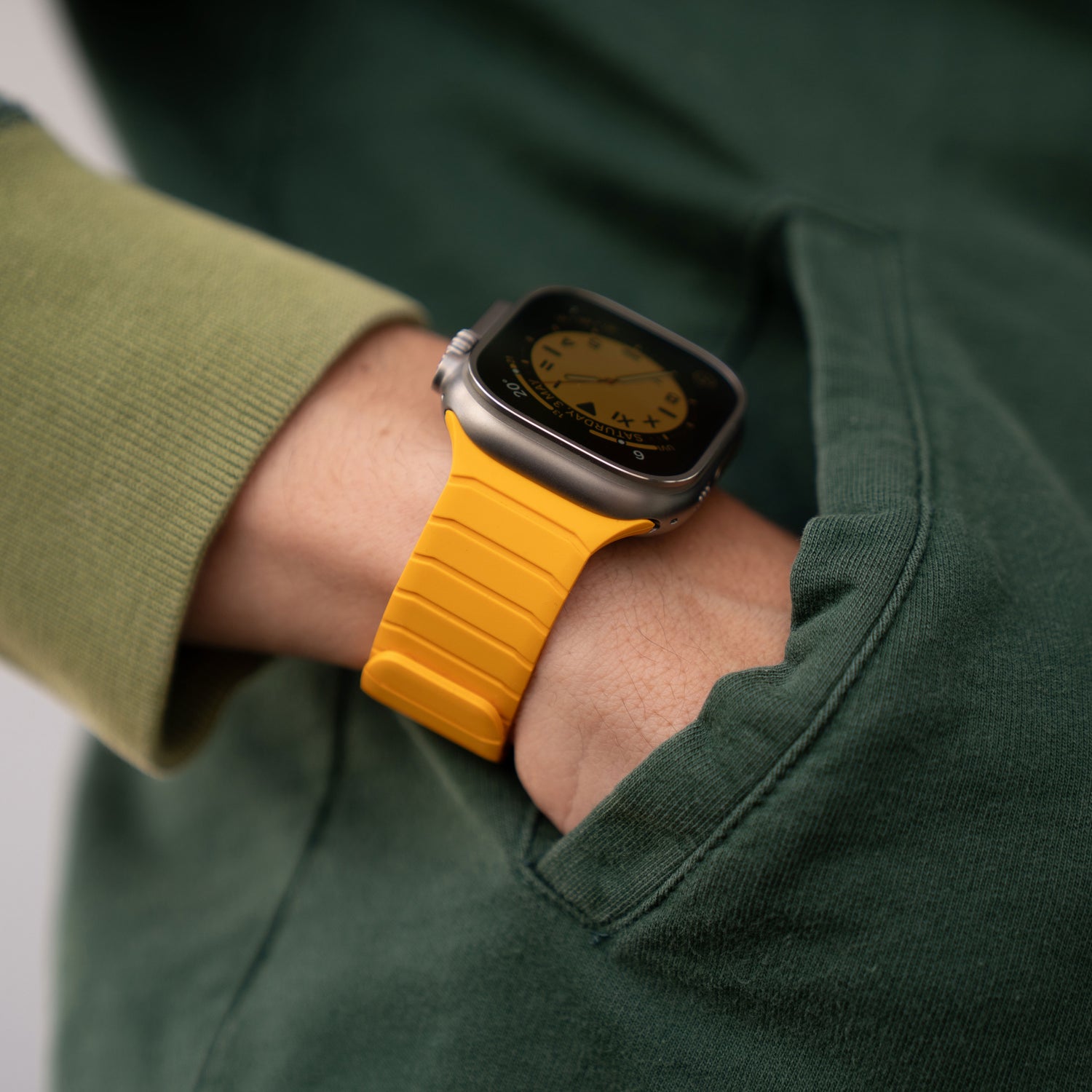 Eine Person in einem grünen Sweatshirt hat die Hand in der Tasche und trägt ein magnetisches Apple Watch Armband „MagFlow“ mit gelbem Silikonband. Das analoge Zifferblatt ist auf dem Display deutlich zu erkennen.