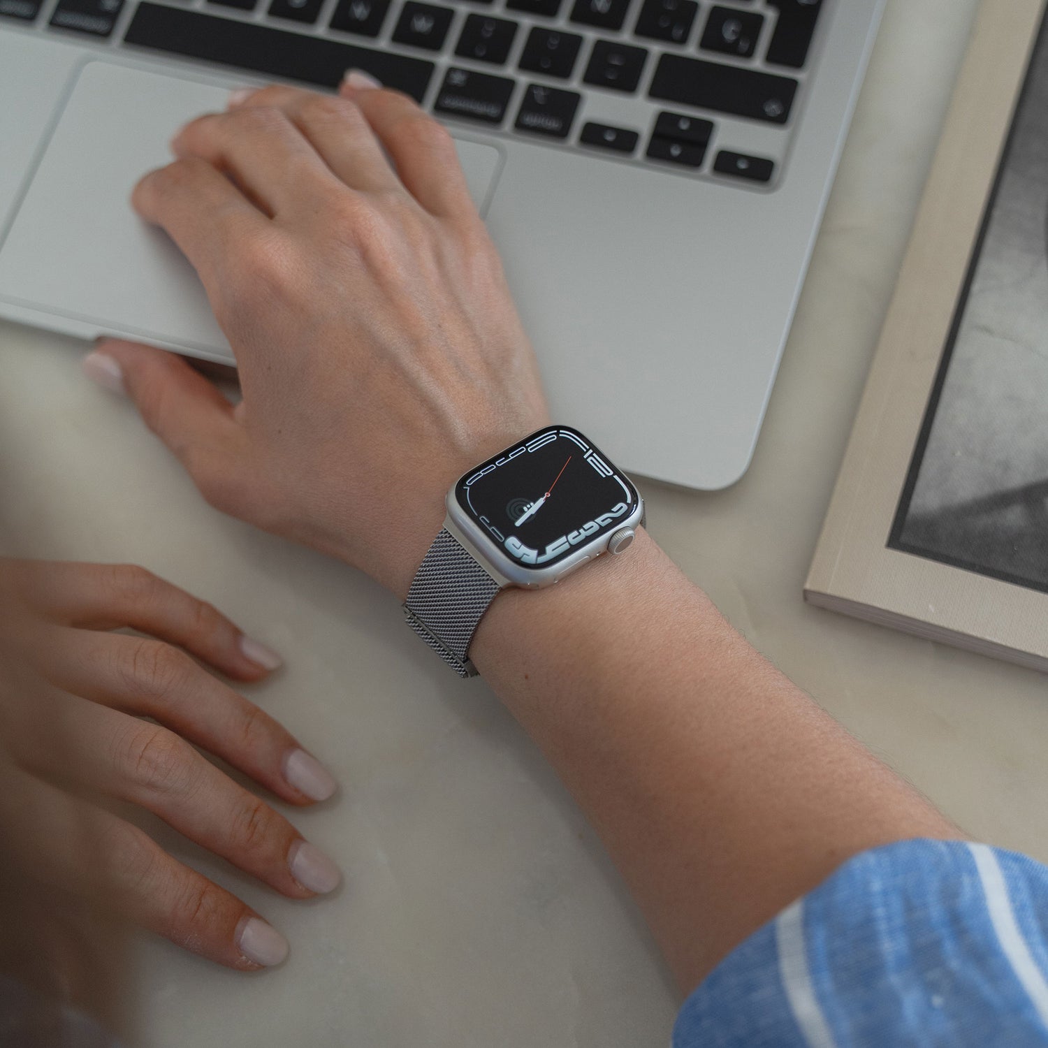 Jemand, der ein arktisband Apple Watch Milanaise Loop Armband von arktisband trägt, tippt auf einer Laptoptastatur, während daneben ein geschlossenes Buch auf der Marmoroberfläche liegt.