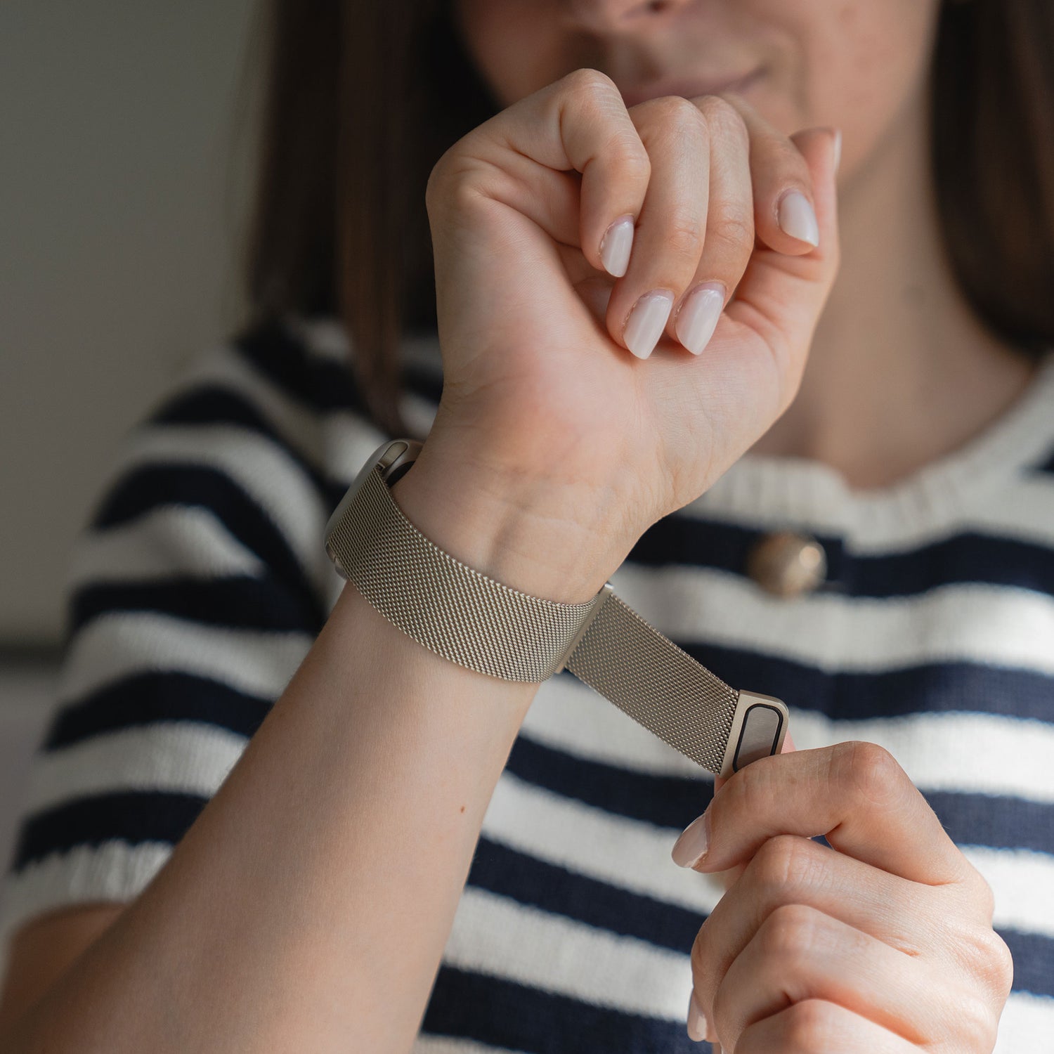 Eine Person in einem marineblau-weiß gestreiften Hemd befestigt ein Arktisband Apple Watch Milanaise Loop Armband um ihr Handgelenk, wobei nur der untere Teil ihres Gesichts und ihrer Hände sichtbar ist.
