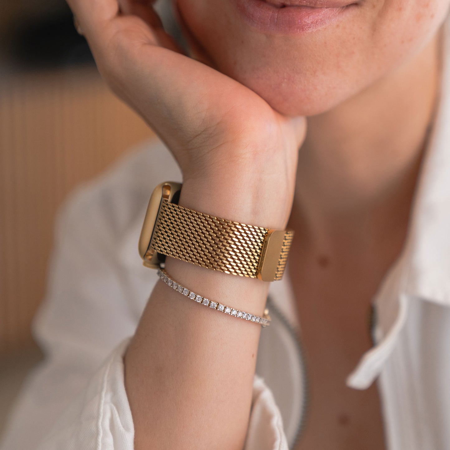 Eine Person trägt ein Arktisband Apple Watch Milanaise Armband „Bold“ in Gold und ein zartes Diamantarmband und stützt ihr Kinn auf ihre Hand.