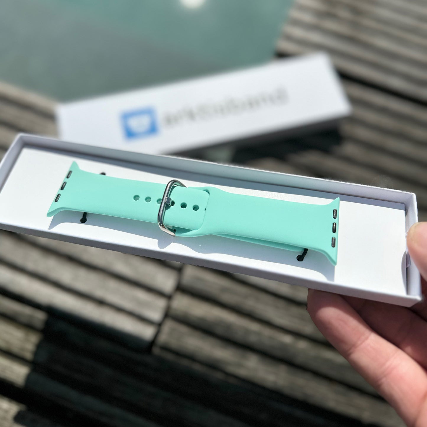 Eine Hand hält eine Schachtel mit dem arktisband Apple Watch Silikonarmband "Aqua" in Mintgrün. Holzbretter und ein verschwommenes weißes Schild mit blauem Text bilden den Hintergrund und heben dieses sportliche arktisband Accessoire hervor.