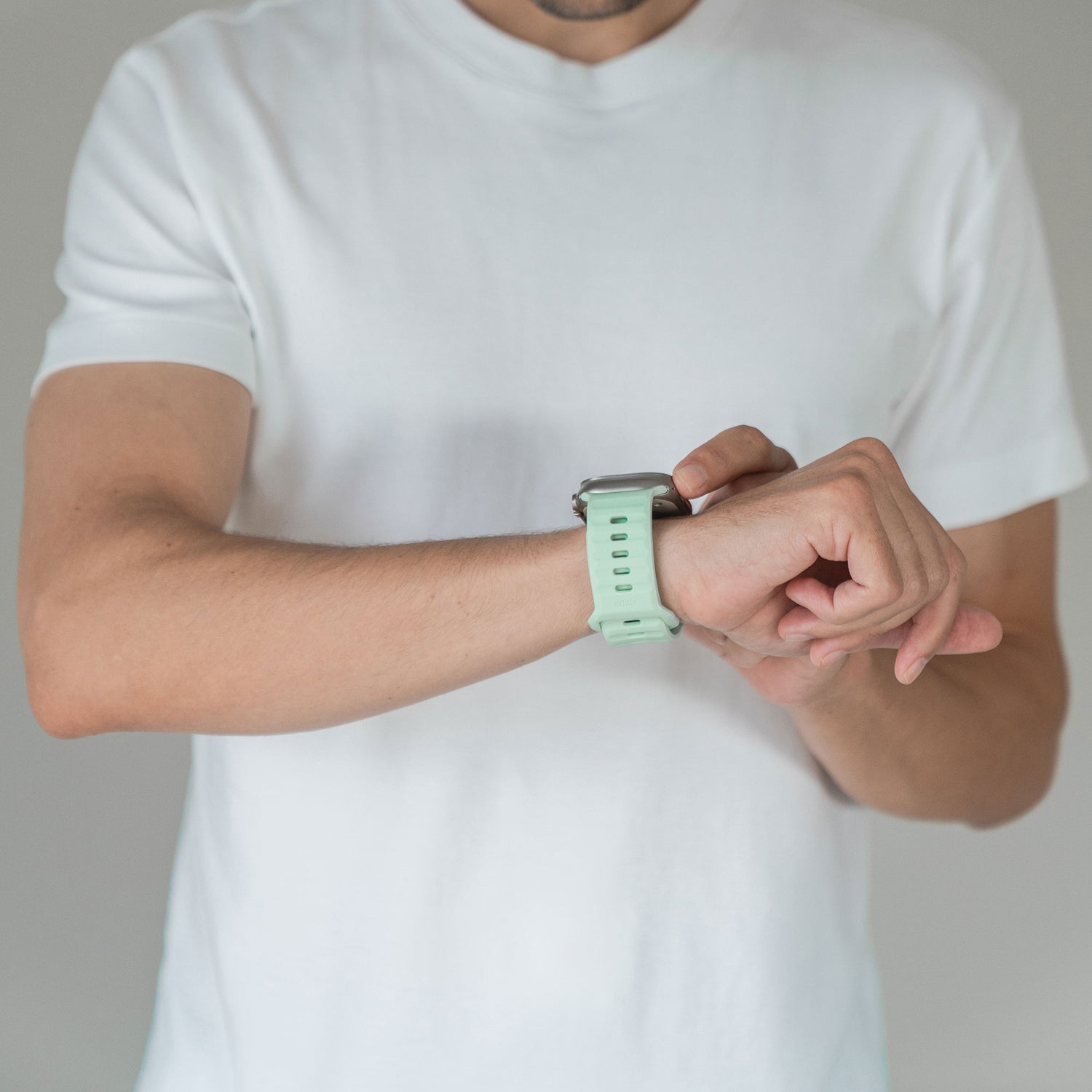 Eine Person in einem schlichten weißen T-Shirt checkt die Uhrzeit auf ihrer Apple Watch mit einem hellgrünen Arktisband Apple Watch Silikonarmband „Active“ und sorgt so für einen sportlichen Look. Das Bild ist so zugeschnitten, dass nur Oberkörper und Arme zu sehen sind.