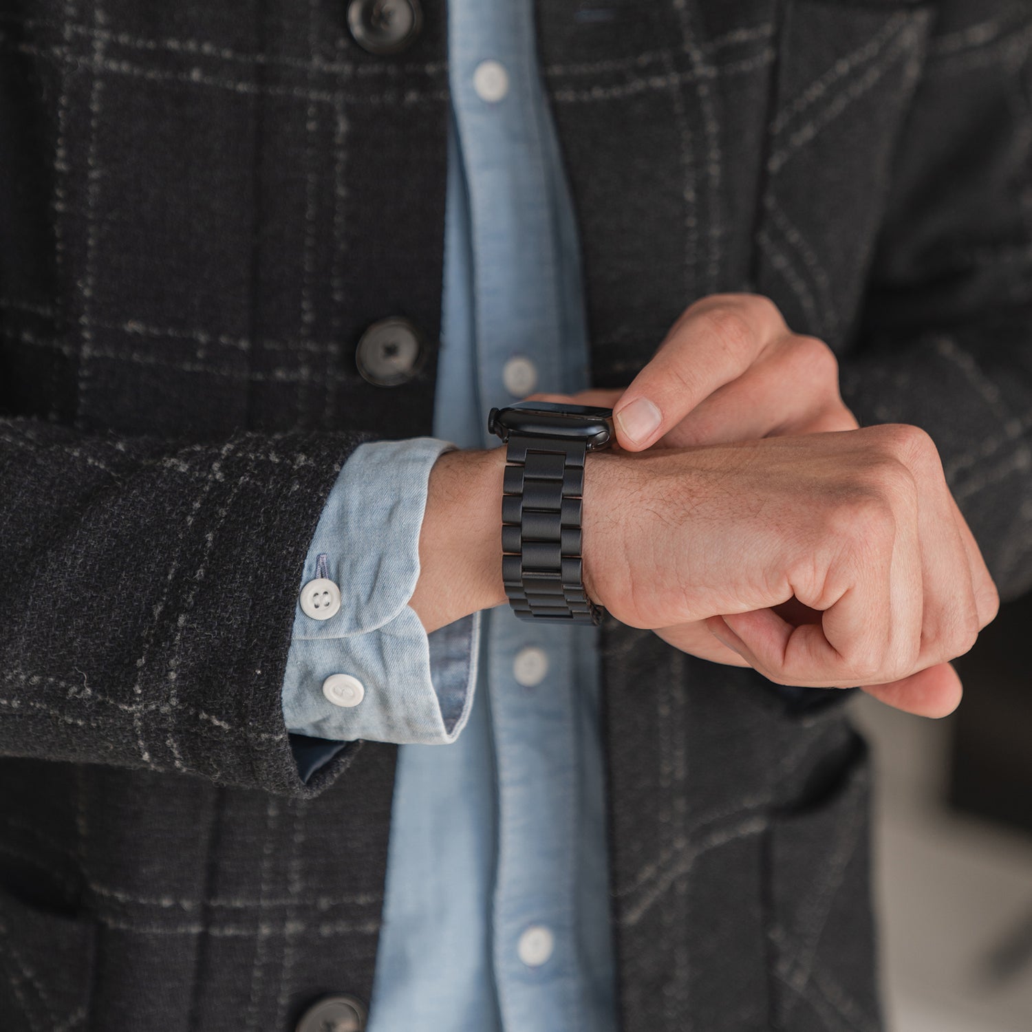 Eine Person in einem dunkelkarierten Blazer und einem hellblauen Hemd rückt das Arktisband Apple Watch Gliederarmband „Arctica“ an ihrem linken Handgelenk zurecht. Die Aufnahme konzentriert sich auf die Hände und das Arktisband-Produkt; ihr Gesicht ist nicht zu sehen.