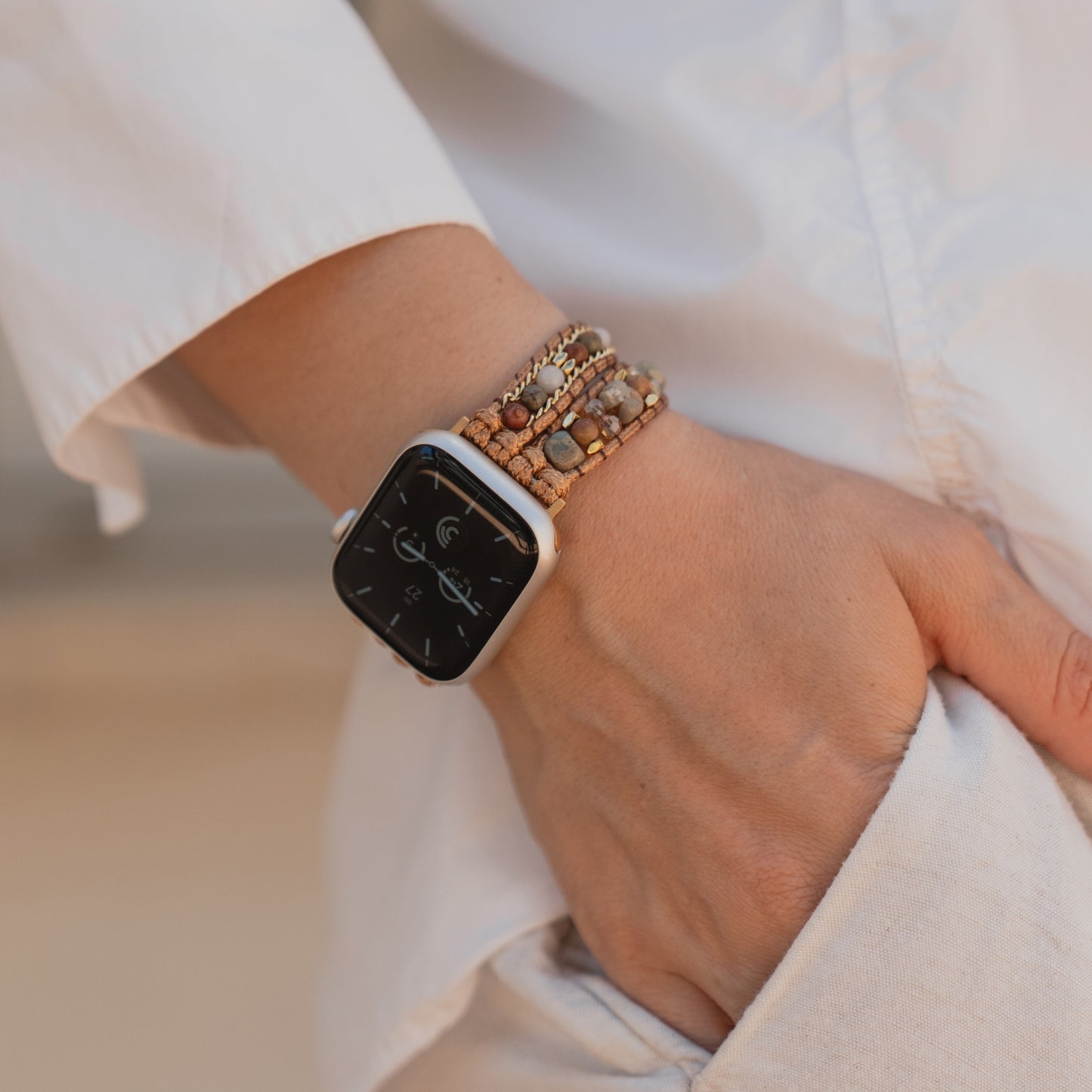 Nahaufnahme einer Hand in der Hosentasche, die das arktisband Apple Watch-Armband „Sunset Spirit“ und ein Perlenarmband über einem weißen Hemd trägt.