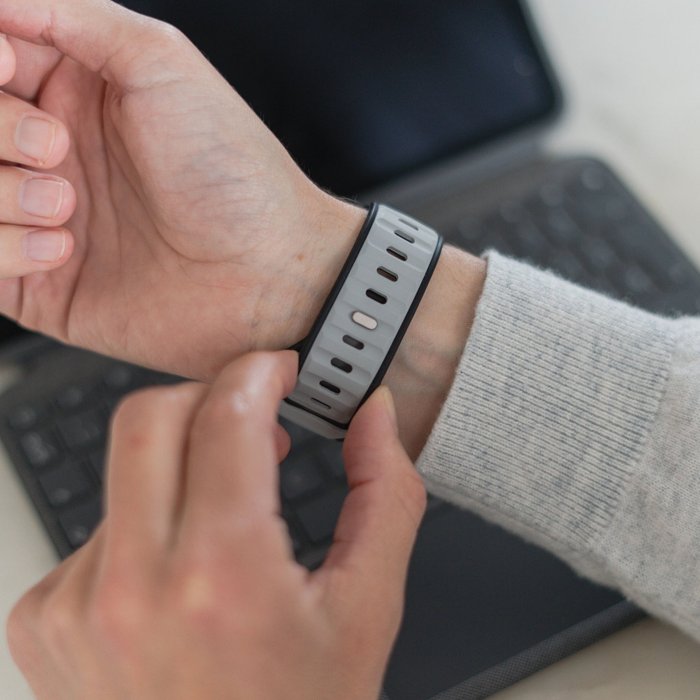 Eine Person befestigt das Arktisband Apple Watch Silikonarmband „Swift“ – ein grau-schwarzes, schweißabsorbierendes und atmungsaktives Band – an ihrem Handgelenk, im Hintergrund ist eine verschwommene Laptoptastatur zu sehen.
