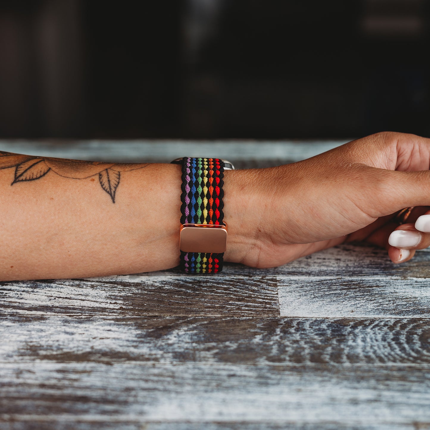Ein Handgelenk mit einem Blättertattoo trägt das arktisband Geflochtene Apple Watch Armband „Casual“ Rainbow Edition von arktisband mit einem farbenfrohen geflochtenen Band, während die Hand auf einer strukturierten Holzoberfläche ruht.